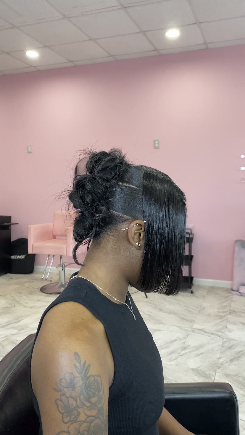 Side view of a woman with short, straight black hair and a tattoo on her shoulder, sitting in a salon.