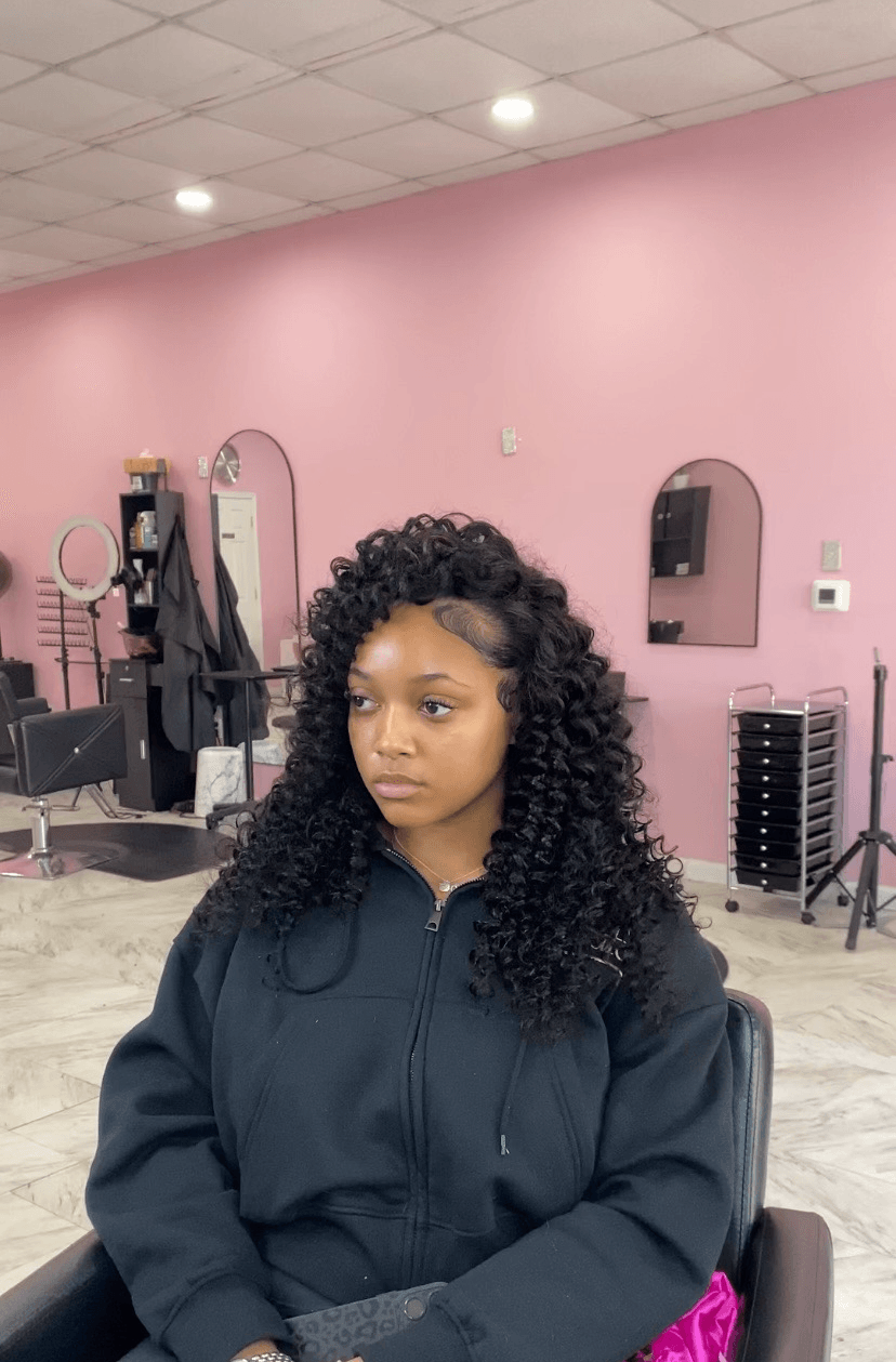 Woman with curly hair sitting in a salon chair, pink wall background, salon equipment visible.