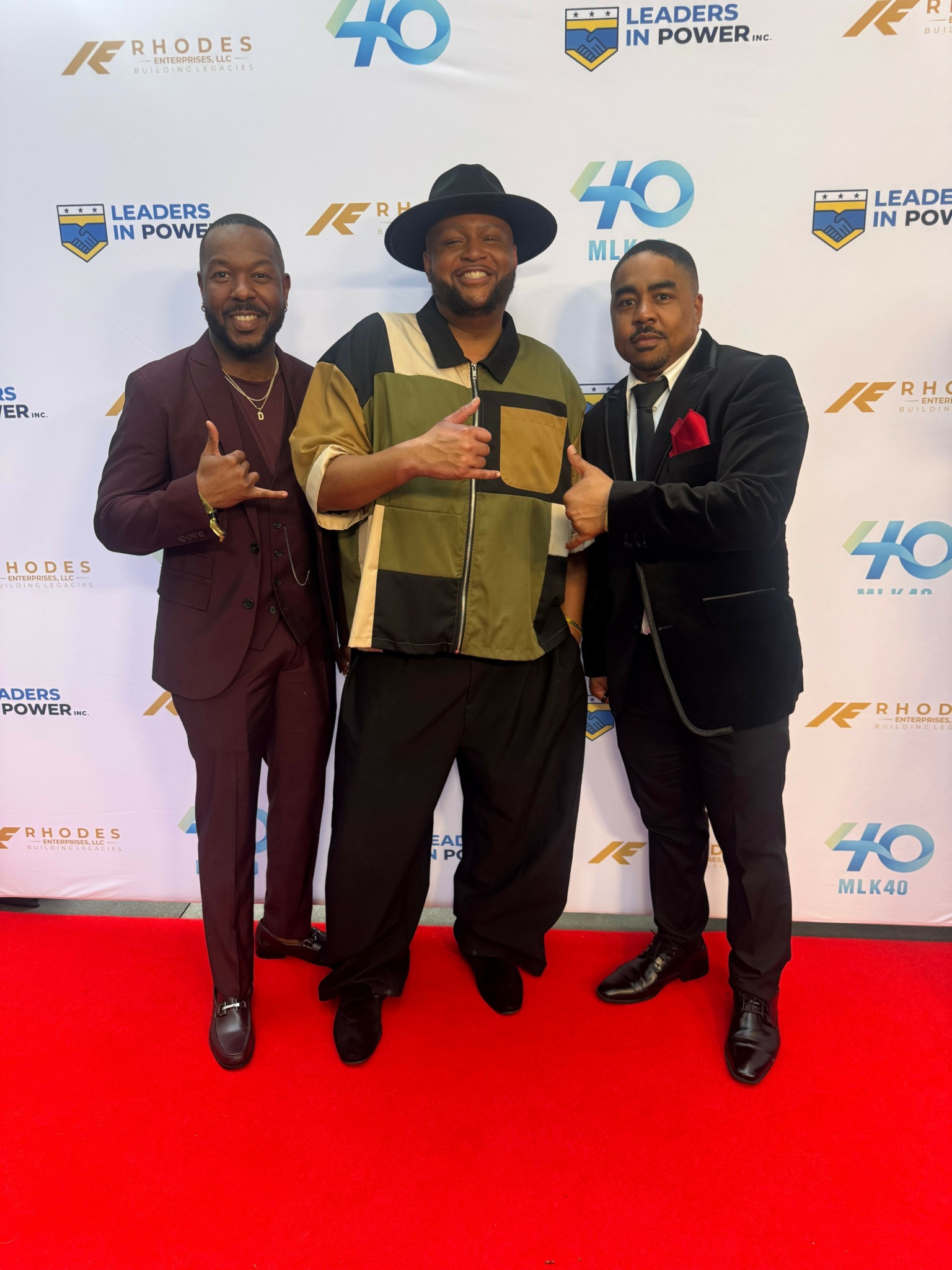 Three people standing on a red carpet in front of a backdrop with logos, posing for a photo.