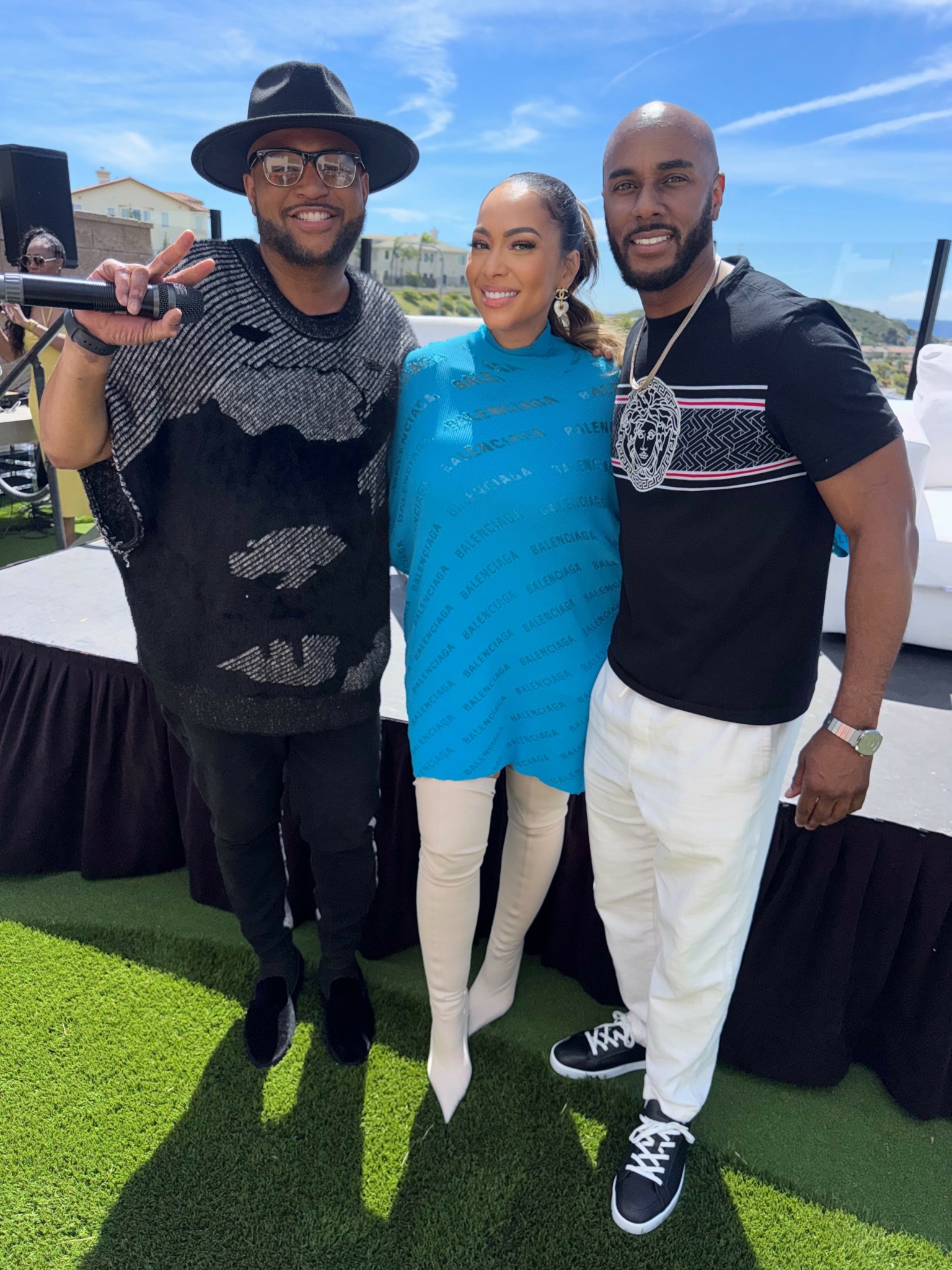 Three people standing outdoors on grass, smiling, with a blue sky and buildings in background.