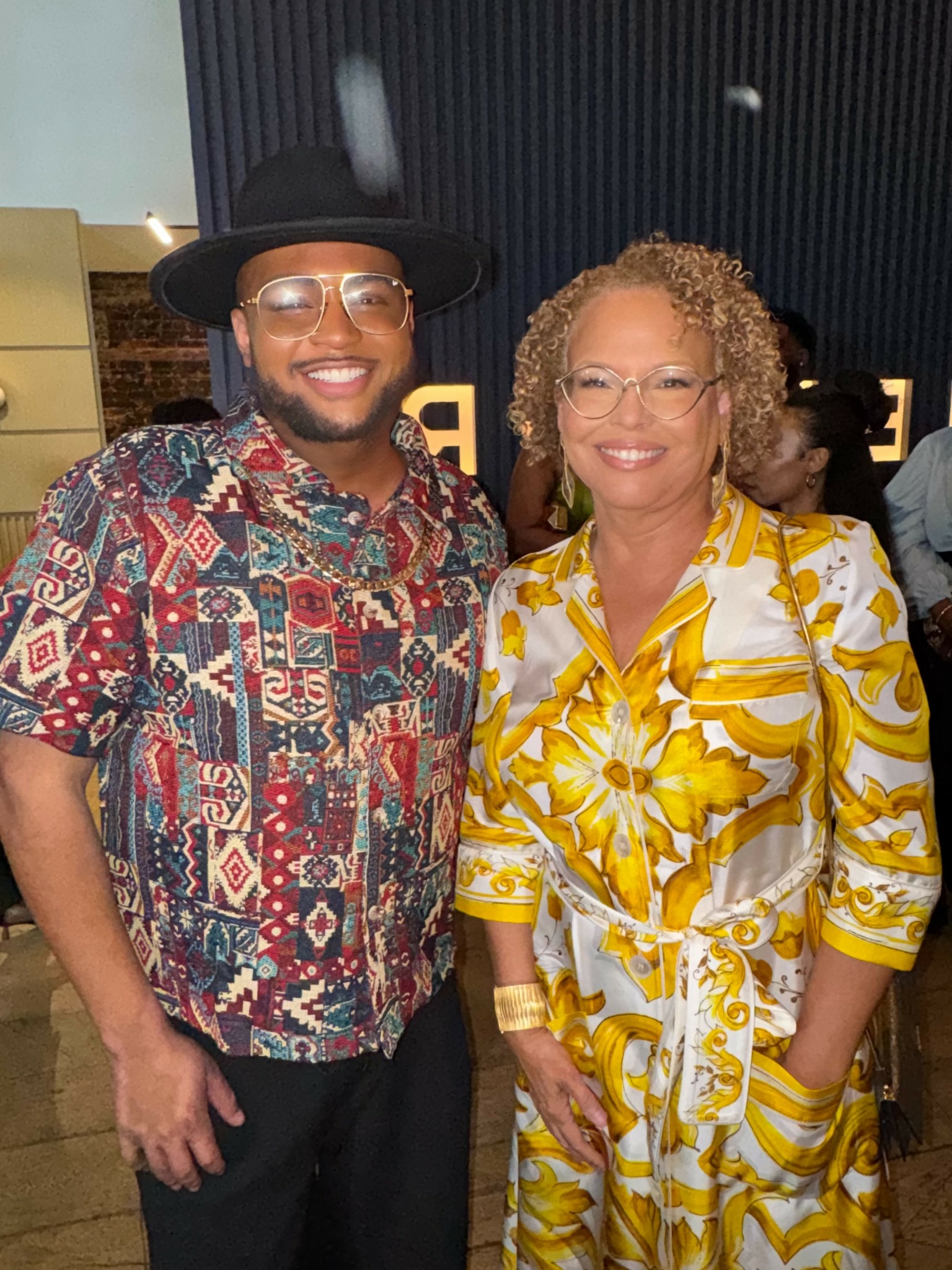 Two smiling people, one wearing a colorful patterned shirt and hat, the other in a yellow and white patterned dress, stand together indoors.