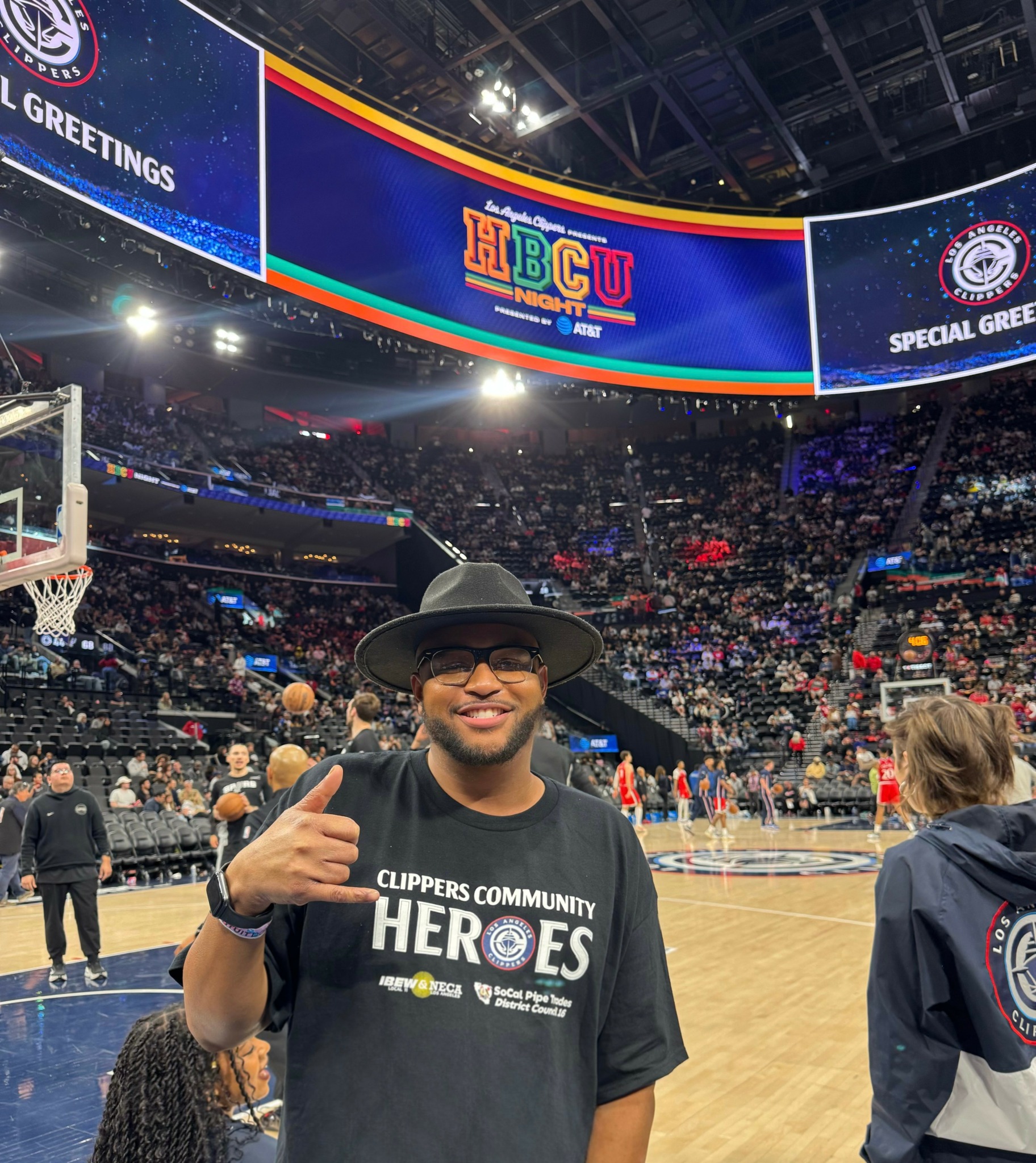 Person wearing a hat and glasses giving a thumbs-up at a basketball game, with a large screen overhead and crowd in background.