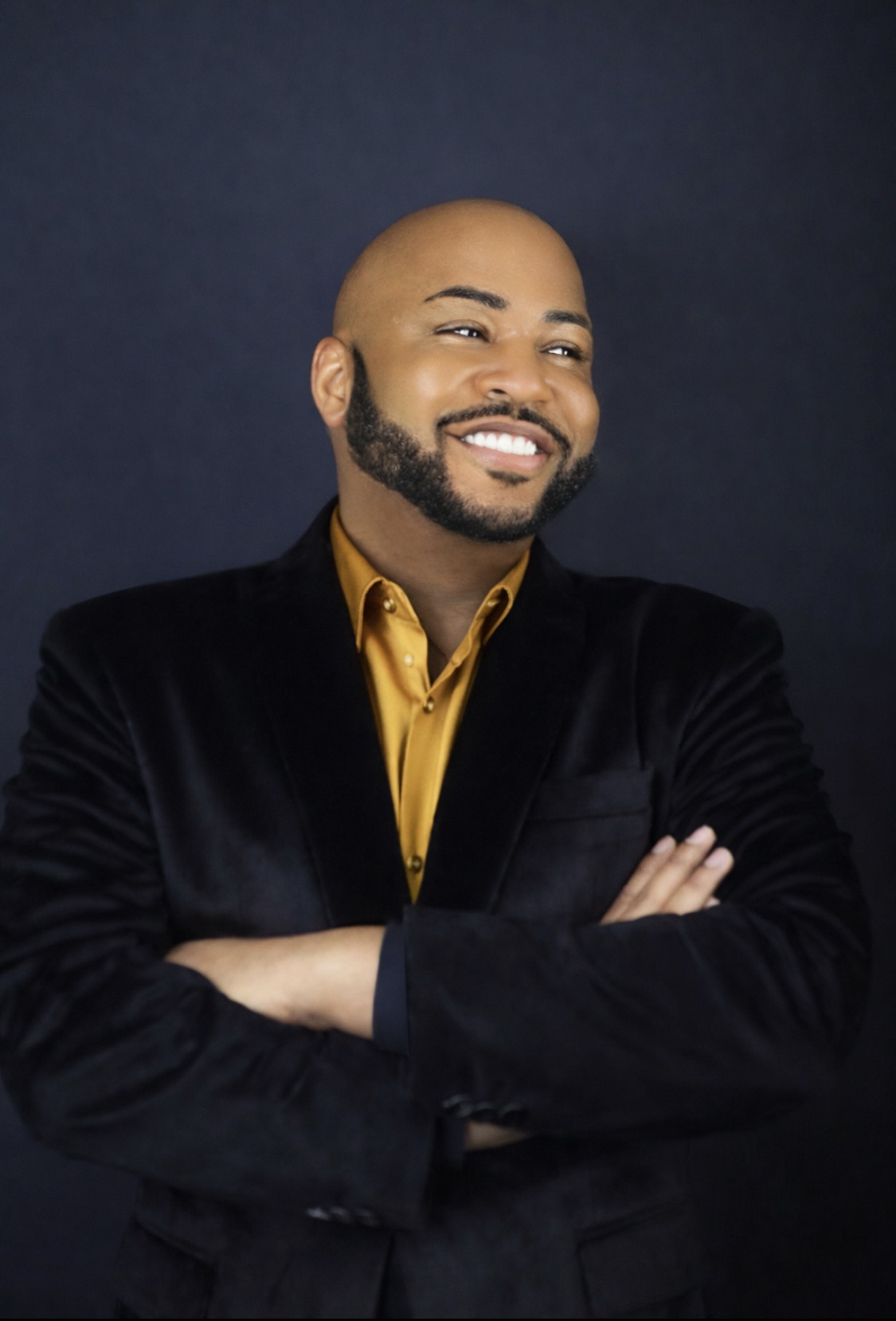 Man with a beard smiling, wearing a black blazer and yellow shirt, arms crossed, dark background.