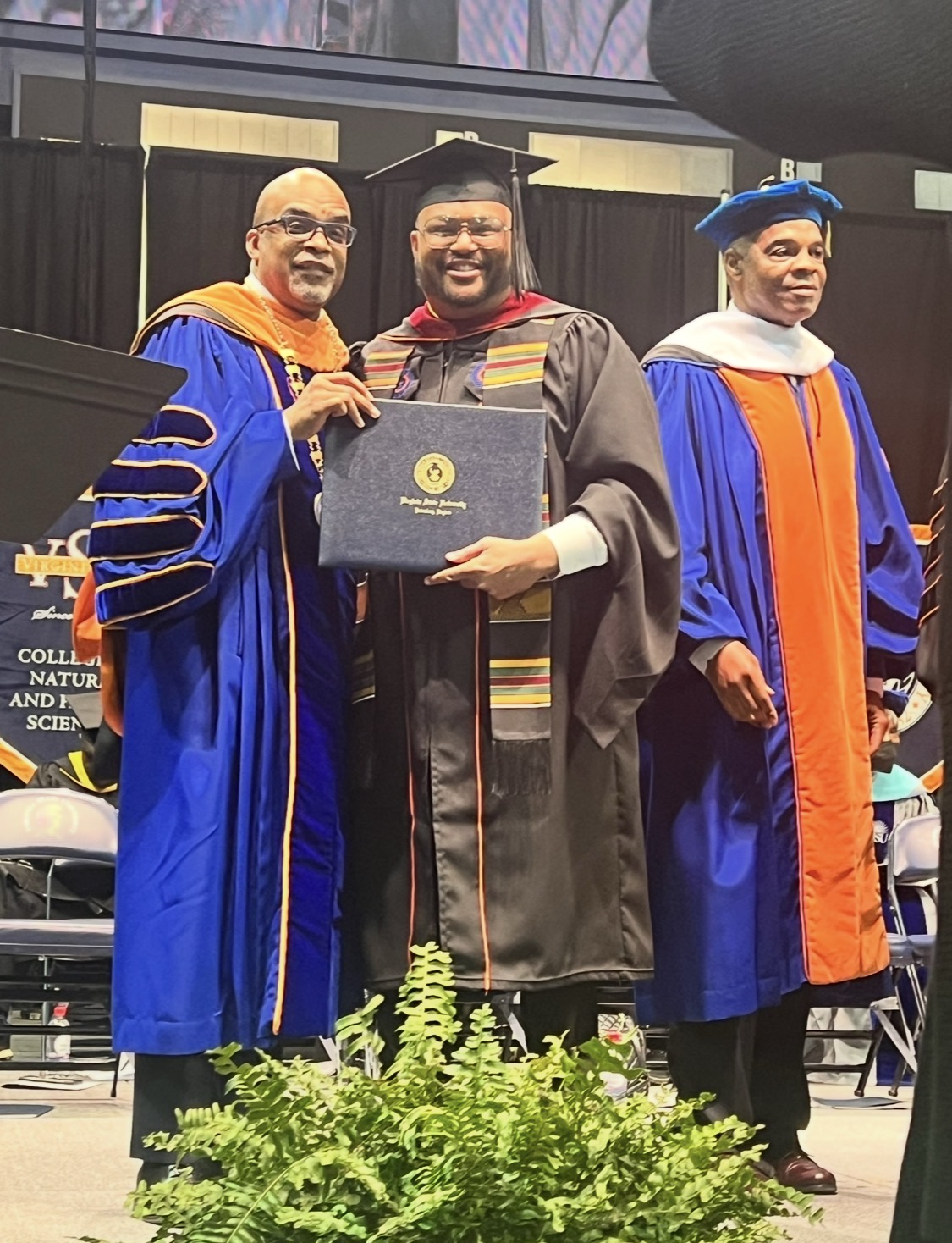 Three people in academic regalia at graduation ceremony, one holding a diploma, smiling, stage background, green plants in foreground.