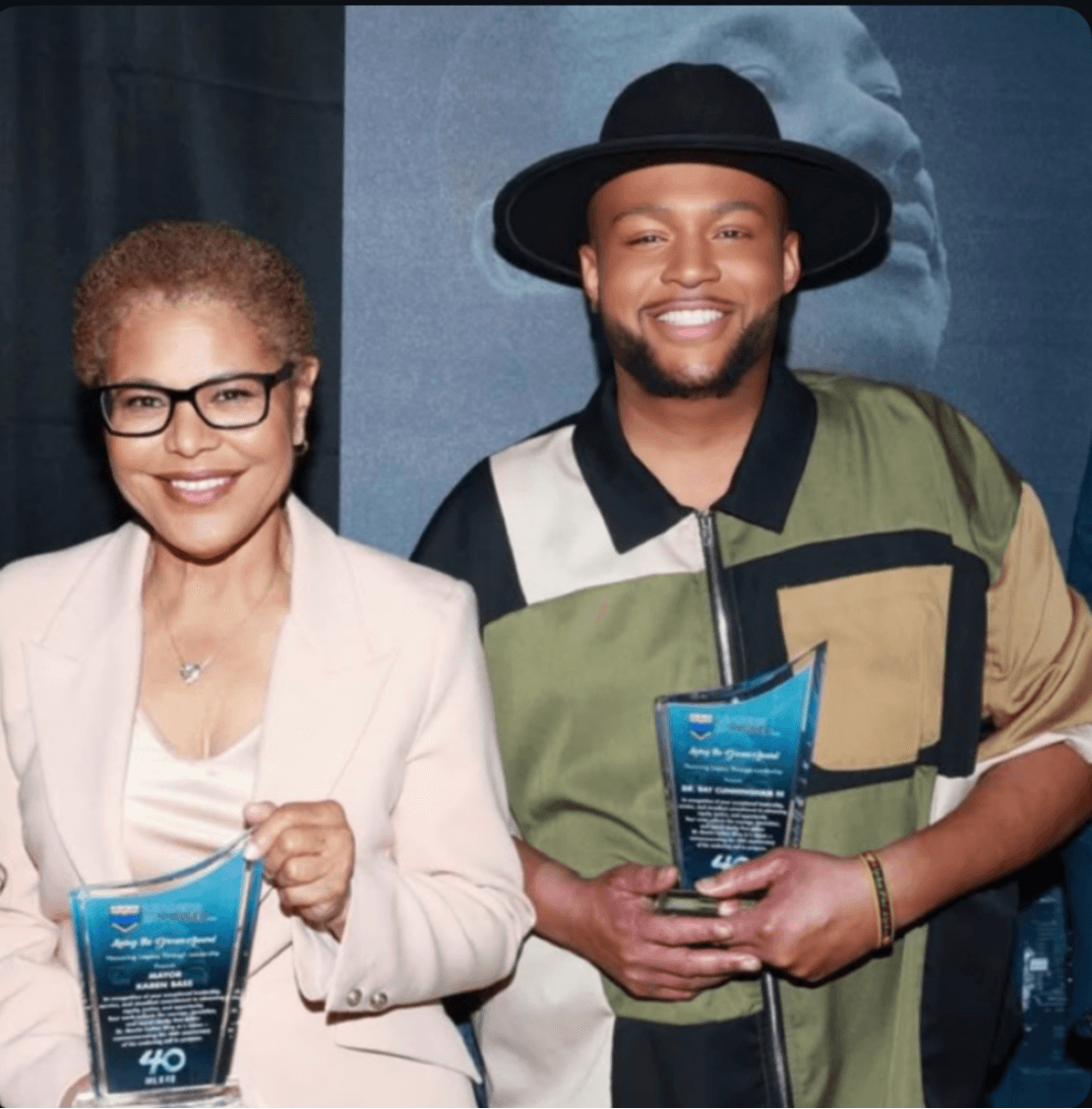 Two people smiling and holding awards, standing indoors with a blue background.