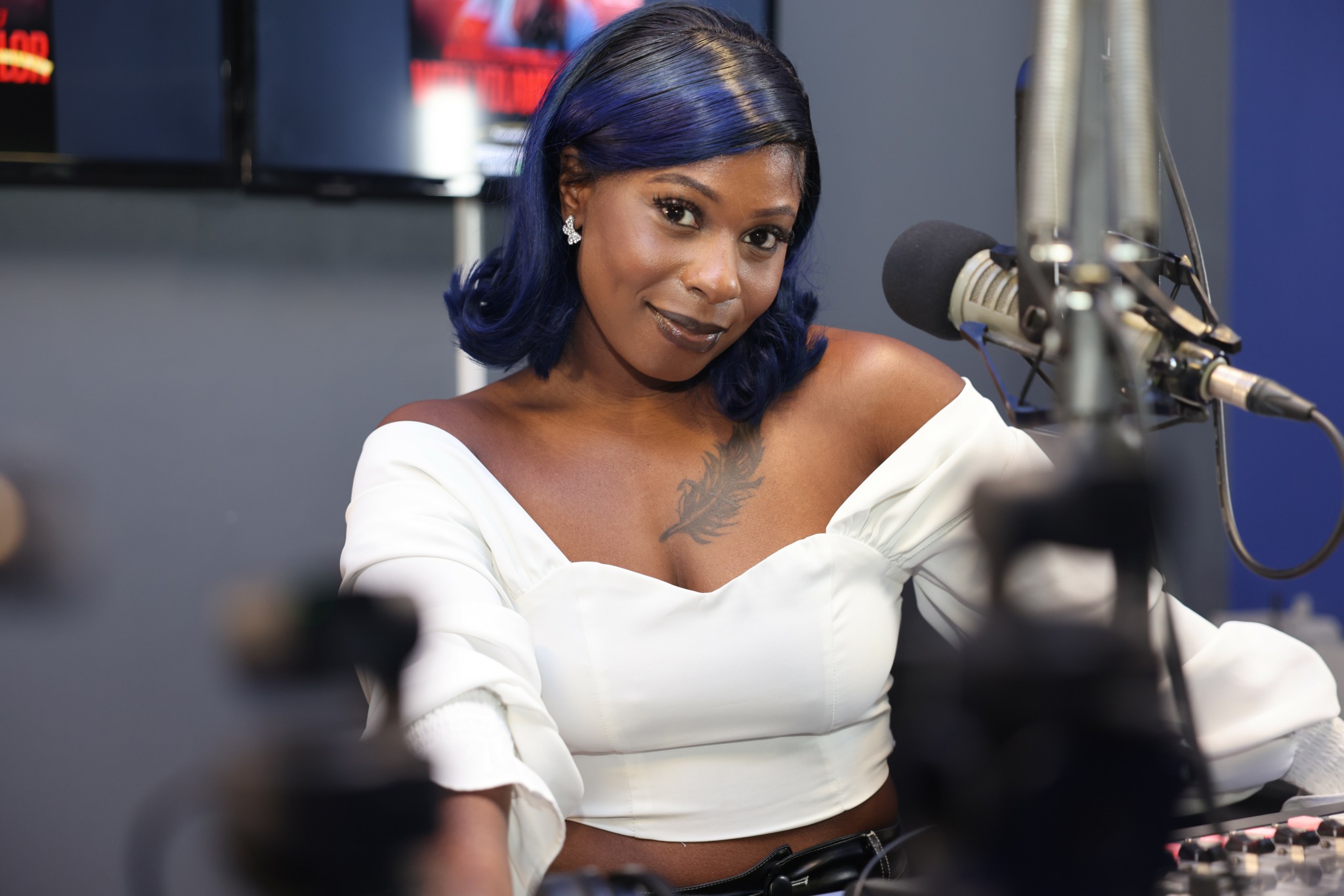 Woman with blue hair speaking into a microphone in a radio studio, wearing a white off-shoulder top.