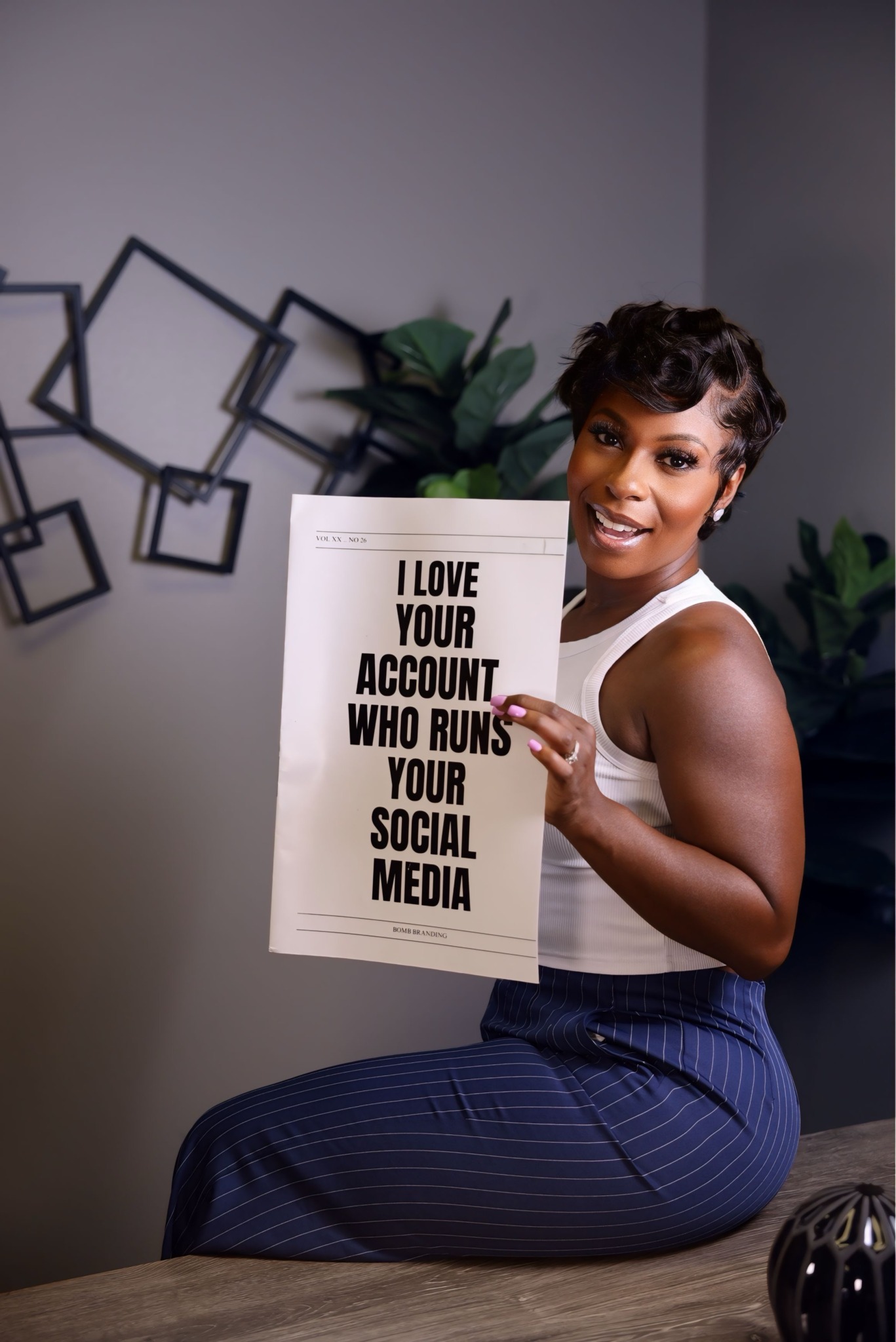 Woman sitting on a table holding a sign with text about social media accounts, smiling at the camera.