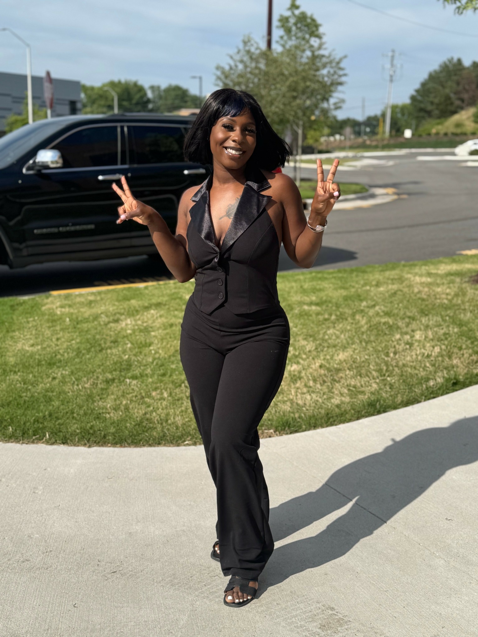 Woman in black jumpsuit smiling and making peace signs outdoors on sidewalk.