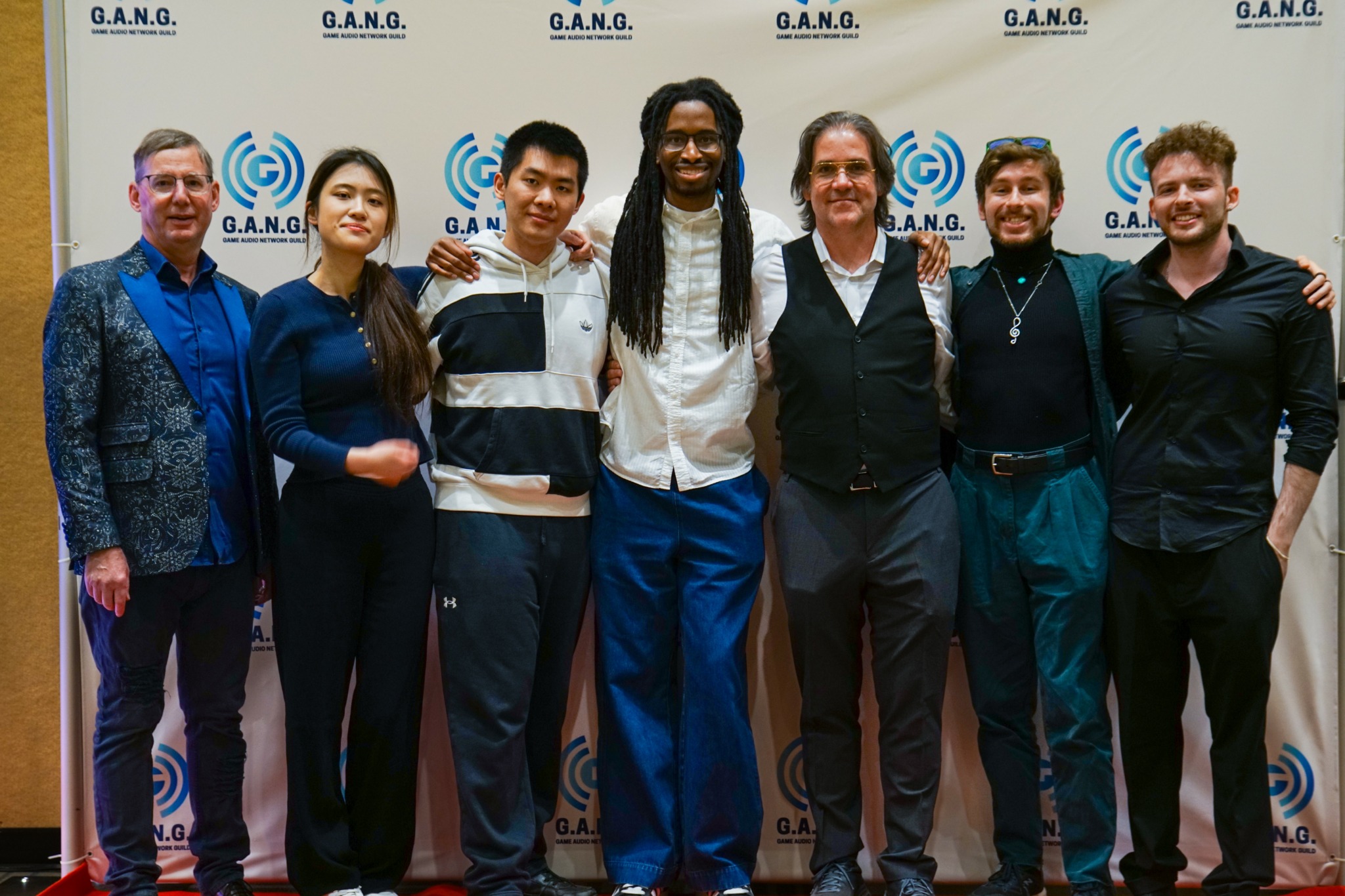 Group of seven people standing together in front of a backdrop with logos, smiling, with arms around each other.