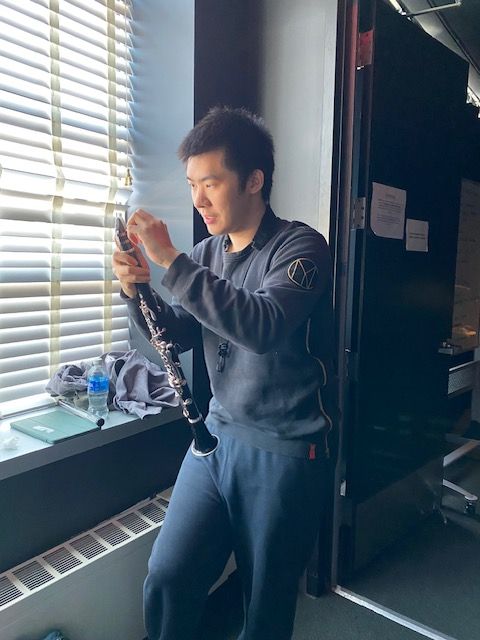 Person with short hair in dark clothing stands near window blinds, holding an object in hand, in a room with a blackboard.