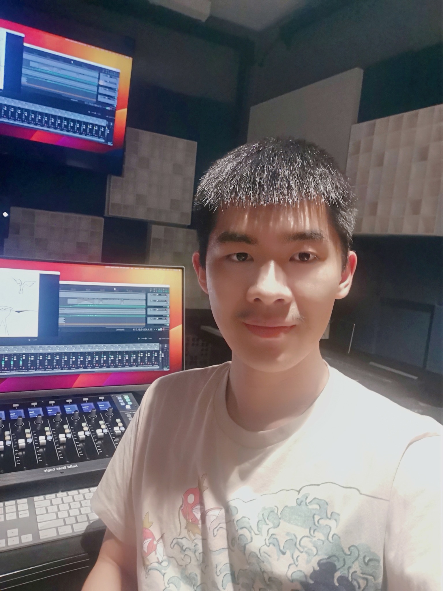Young man with short black hair smiling in a recording studio with audio equipment and screens.