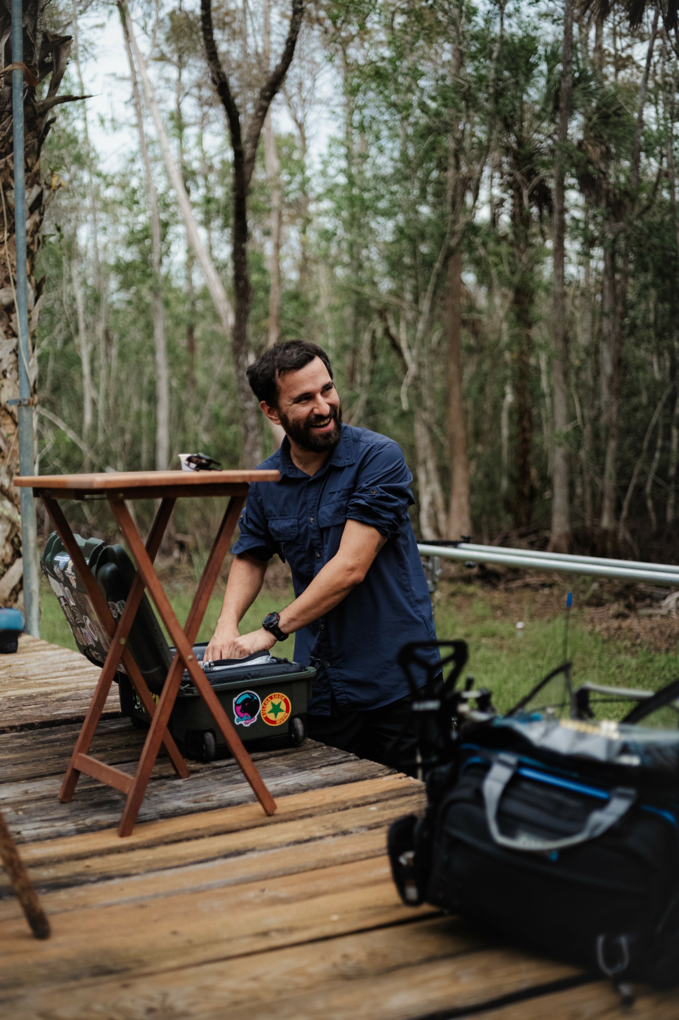 Man with beard smiling, leaning on a wooden table outdoors in a forested area.