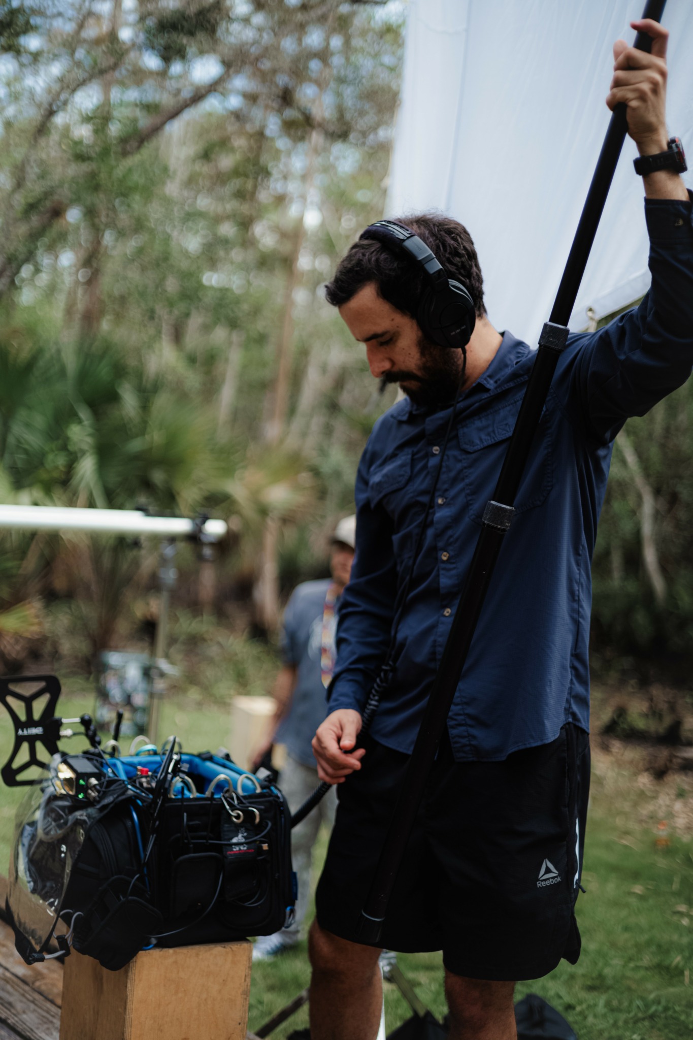 Man with headphones holding a pole outdoors, with a bag on a wooden stand nearby and trees in the background.