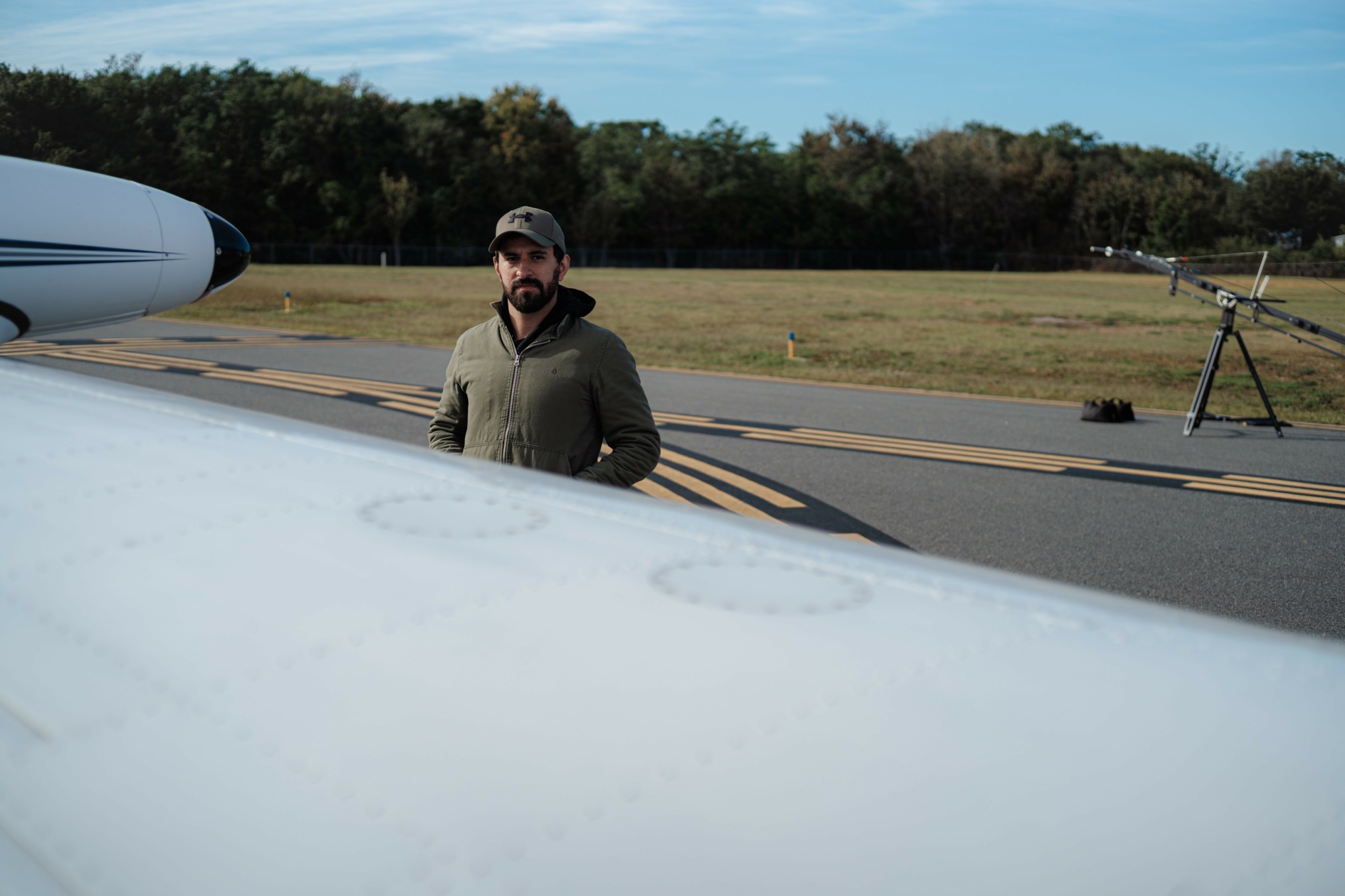 Man standing on tarmac near airplane wing with trees and sky in background.