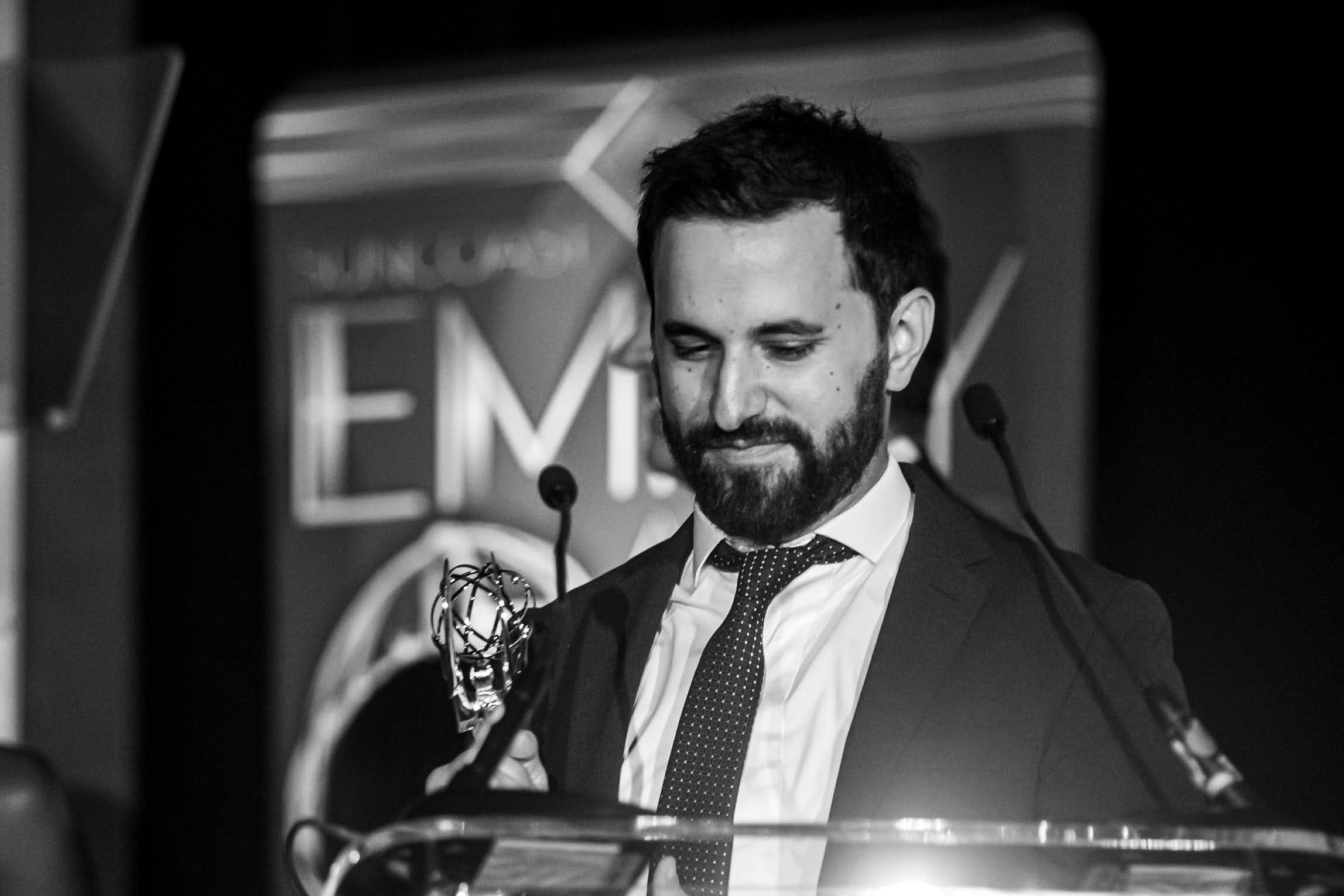 Man with beard in suit and tie speaking at a podium with microphones, background shows a screen with text.