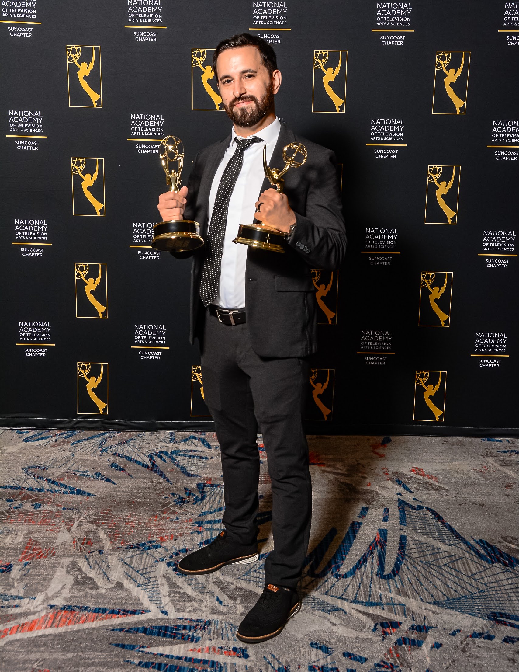 Man in suit holding two awards, standing on patterned carpet, backdrop with award logos.