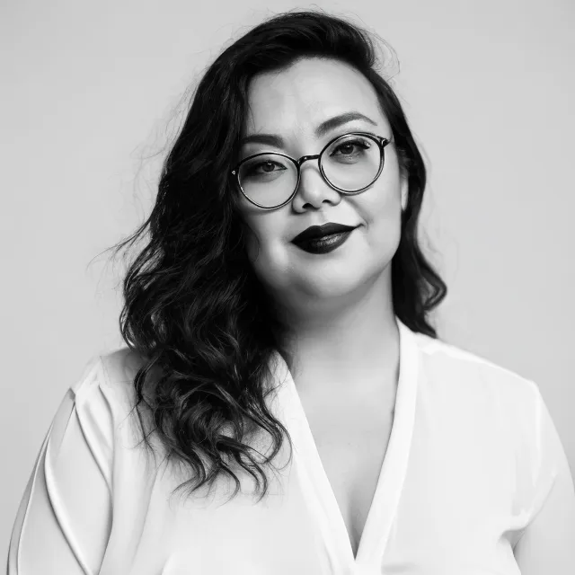 Woman with wavy hair, glasses, and dark lipstick, wearing a white top, looking at camera.