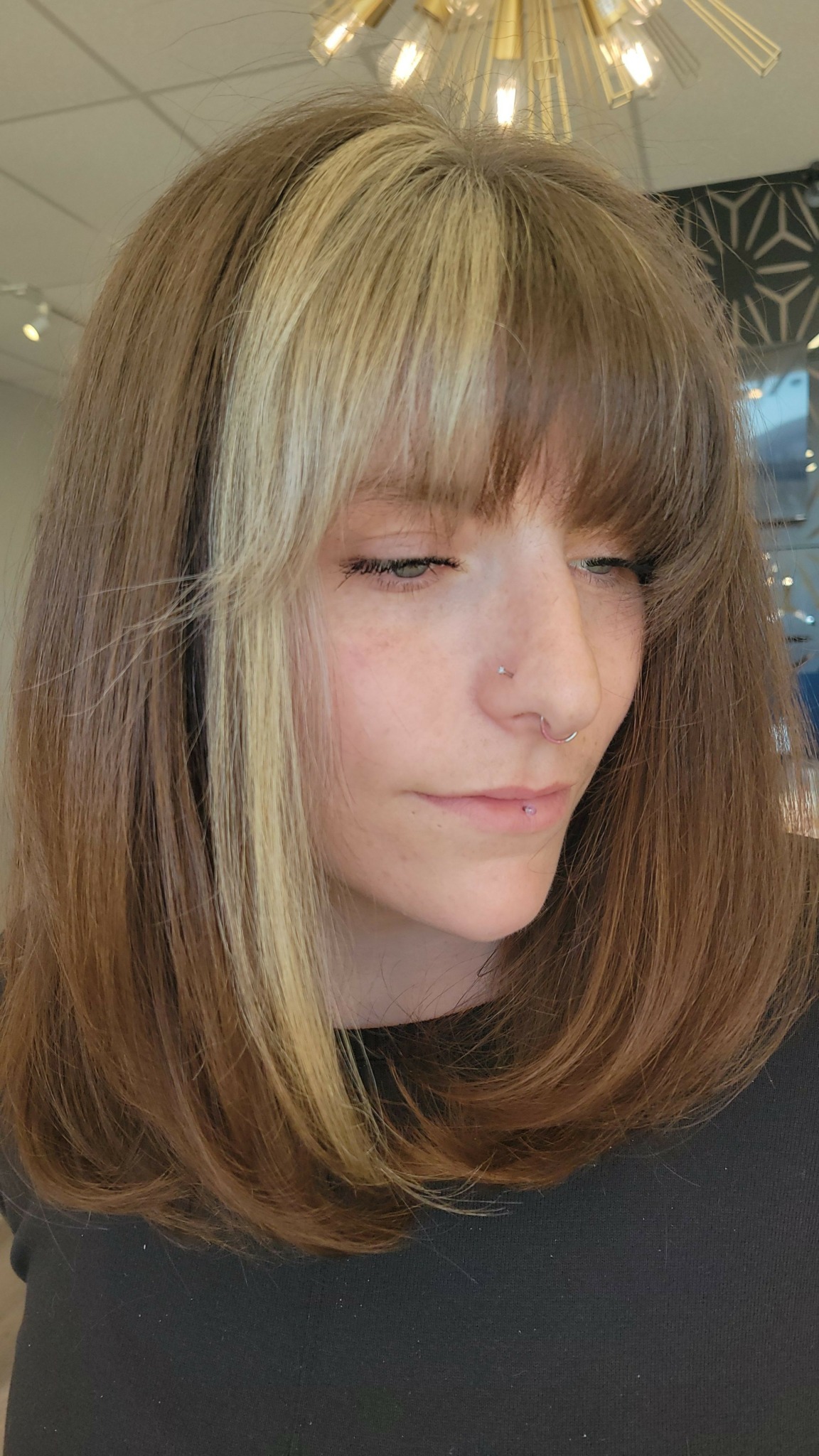 Young woman with long, light brown hair and bangs, eyes closed, indoors with warm lighting.