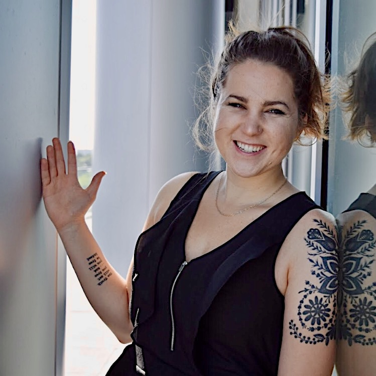 Smiling woman with a tattoo on her arm, wearing a black sleeveless top, standing near a window.