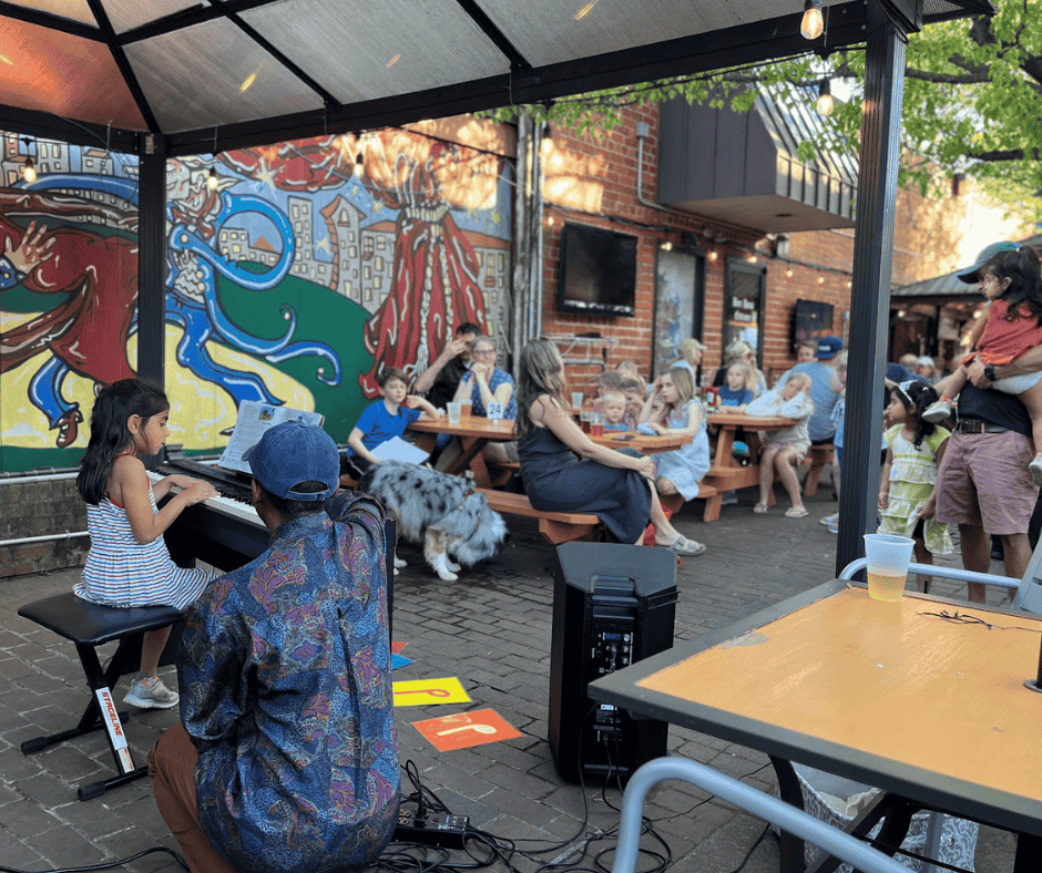 People sitting at tables outdoors, some playing instruments, colorful mural on wall, trees, and a brick building in background.