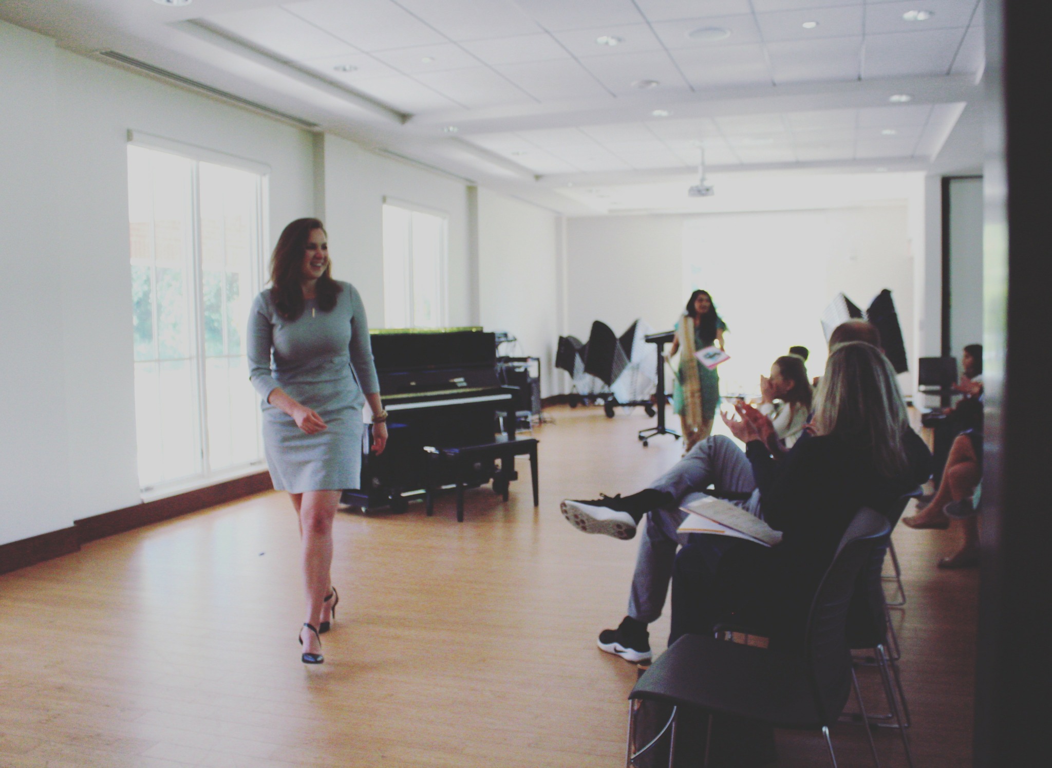 Woman walking in a bright room with people seated and a piano in the background.
