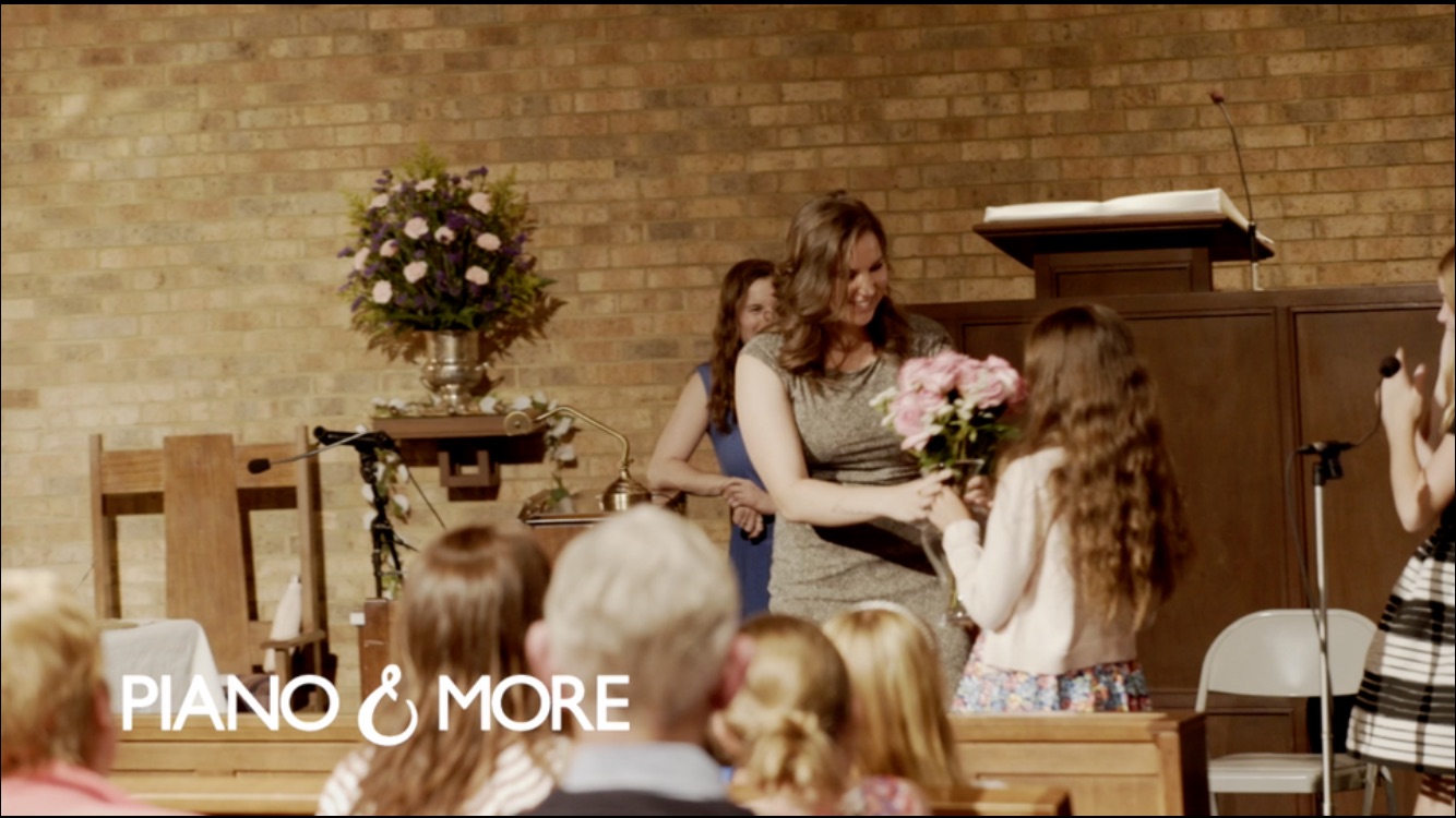 Woman receiving a bouquet from a girl in a room with a brick wall and flowers on a table.