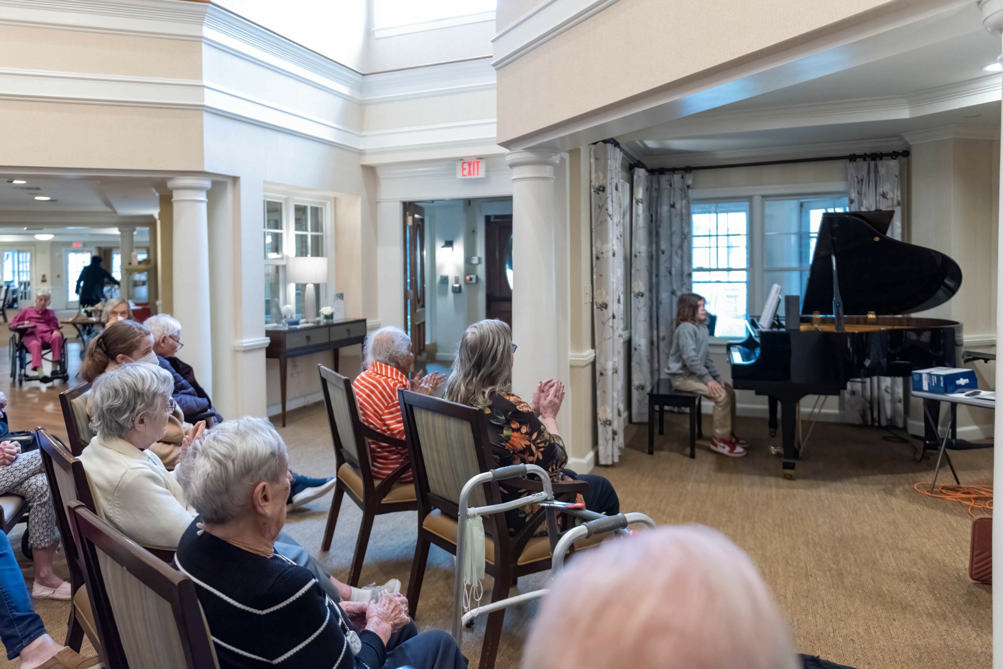 Group of elderly people seated in a room watching a person playing piano near window.