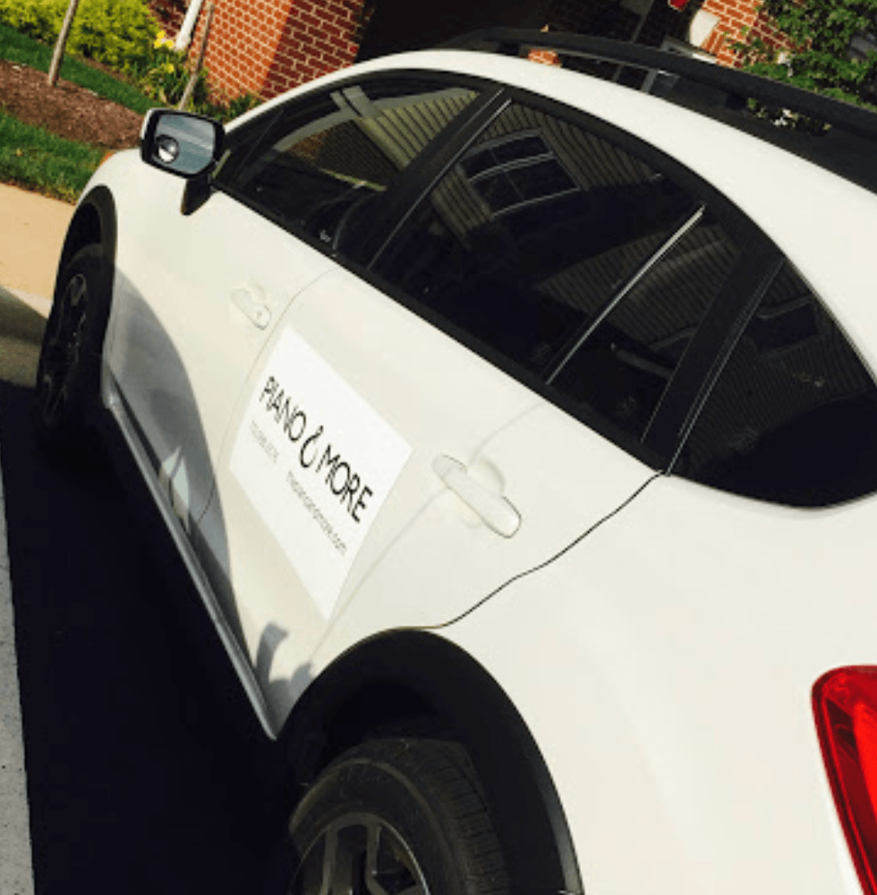 White car parked on street with a sign on the door, brick building and greenery in background.