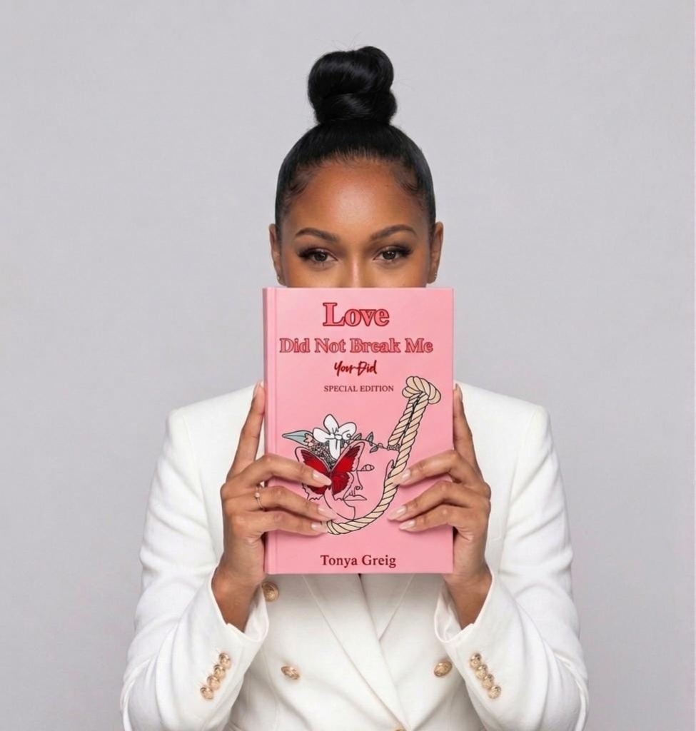 Woman with hair in a bun holds a pink book in front of her face, wearing a white blazer.