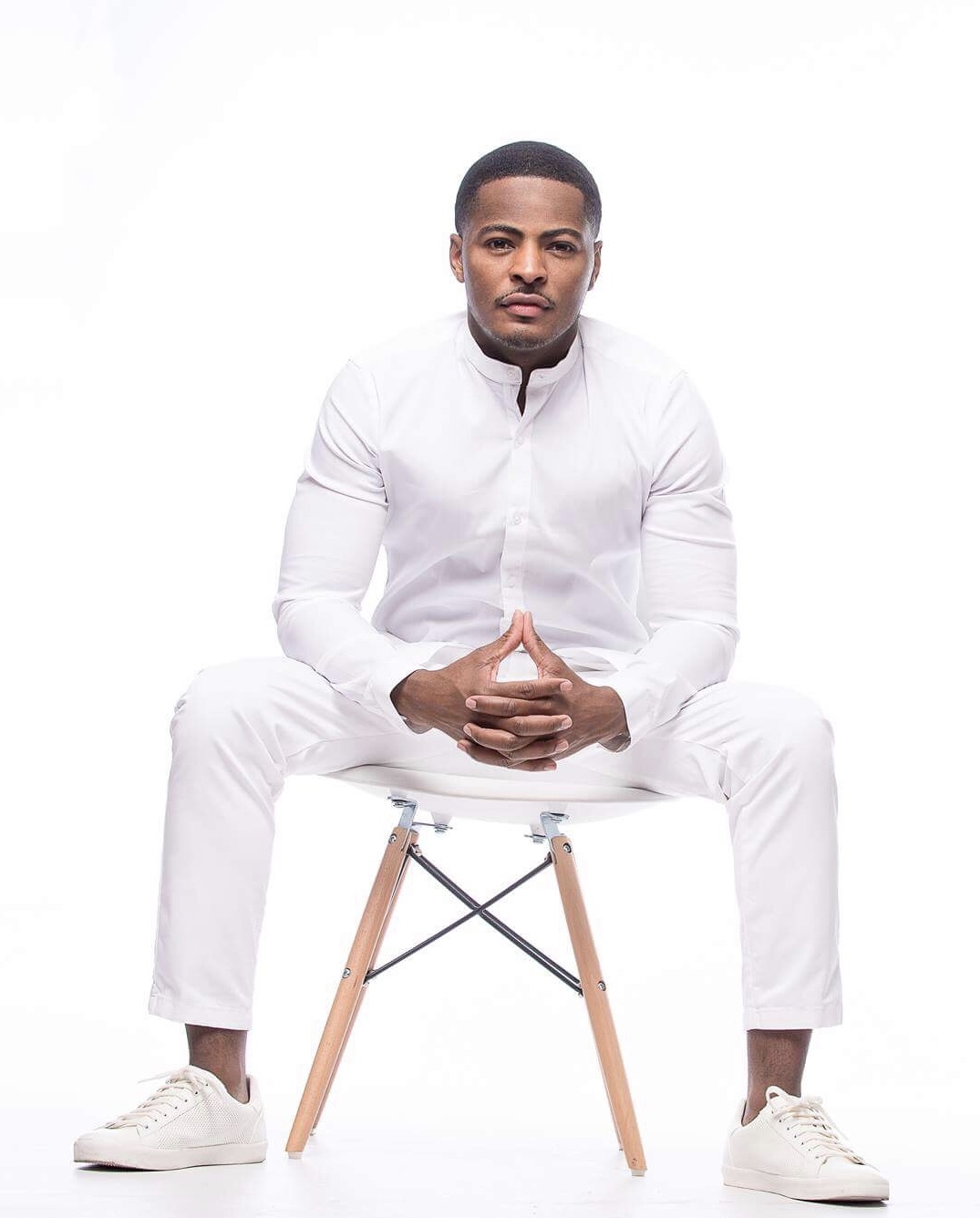 Man sitting on a chair with legs apart, hands clasped, wearing a white outfit, against a white background.