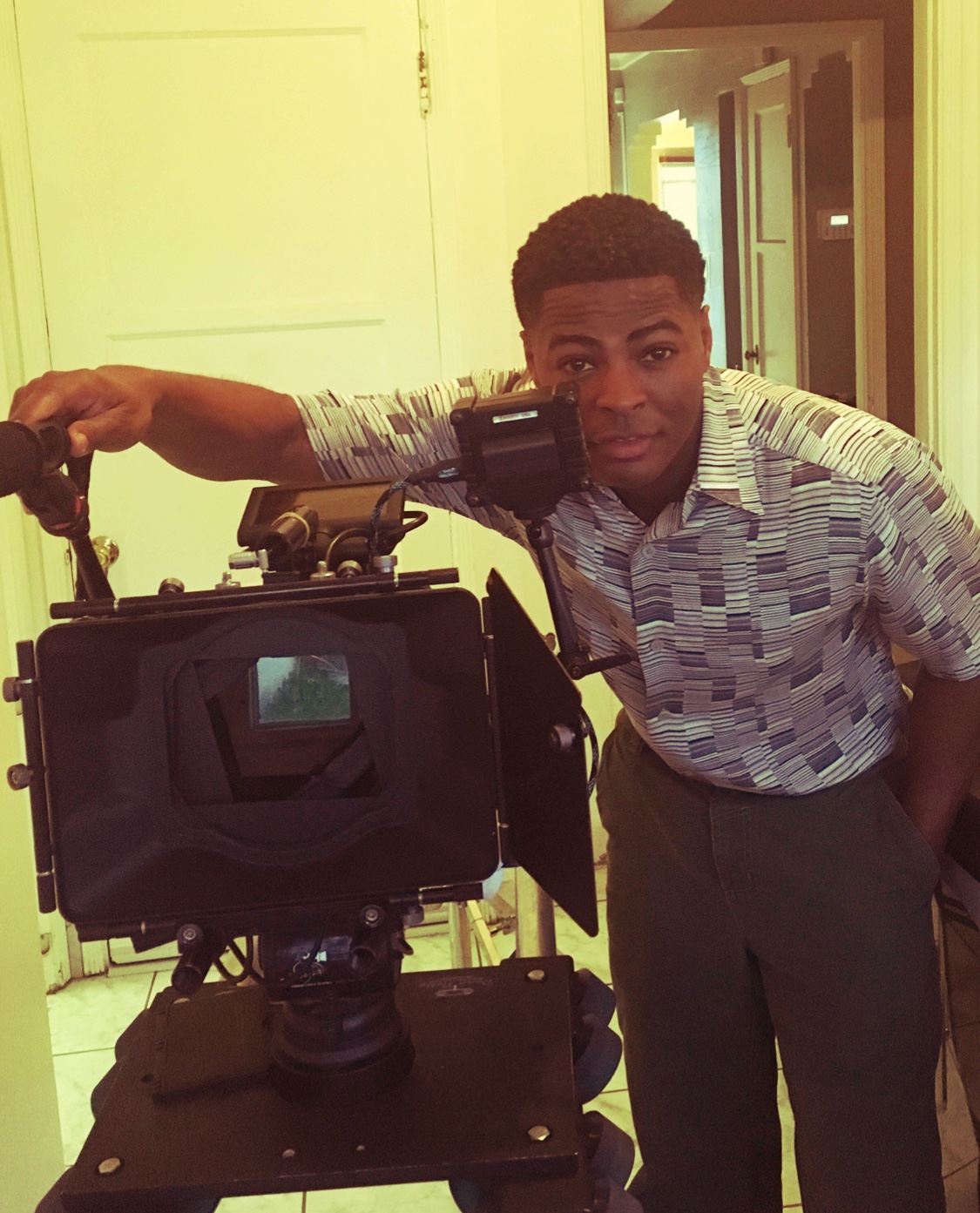Young man with short curly hair leaning on a professional camera, standing in a room with yellow walls and open doors.