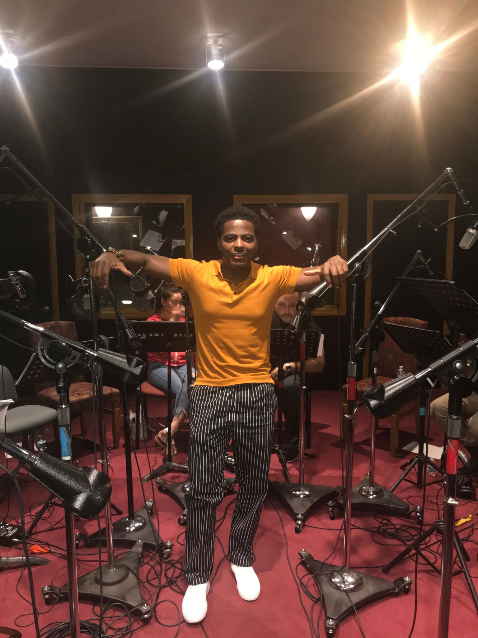 Person in yellow shirt and striped pants holding a microphone in a recording studio with microphones and chairs.