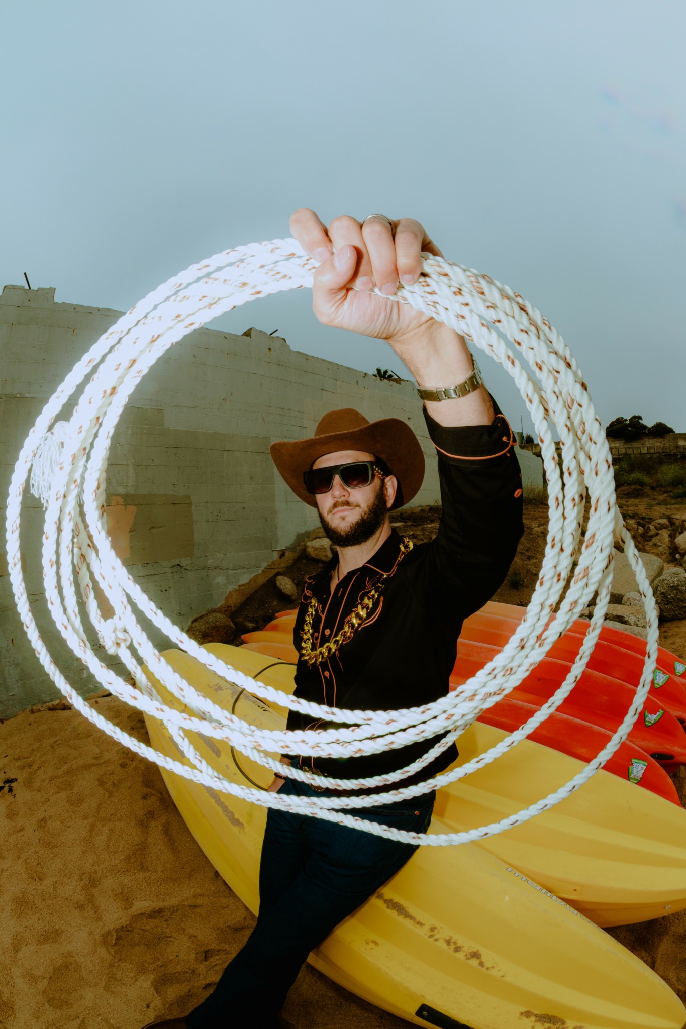 Person wearing sunglasses and a wide-brimmed hat holding a circular light trail in front of yellow kayaks on a sandy beach.