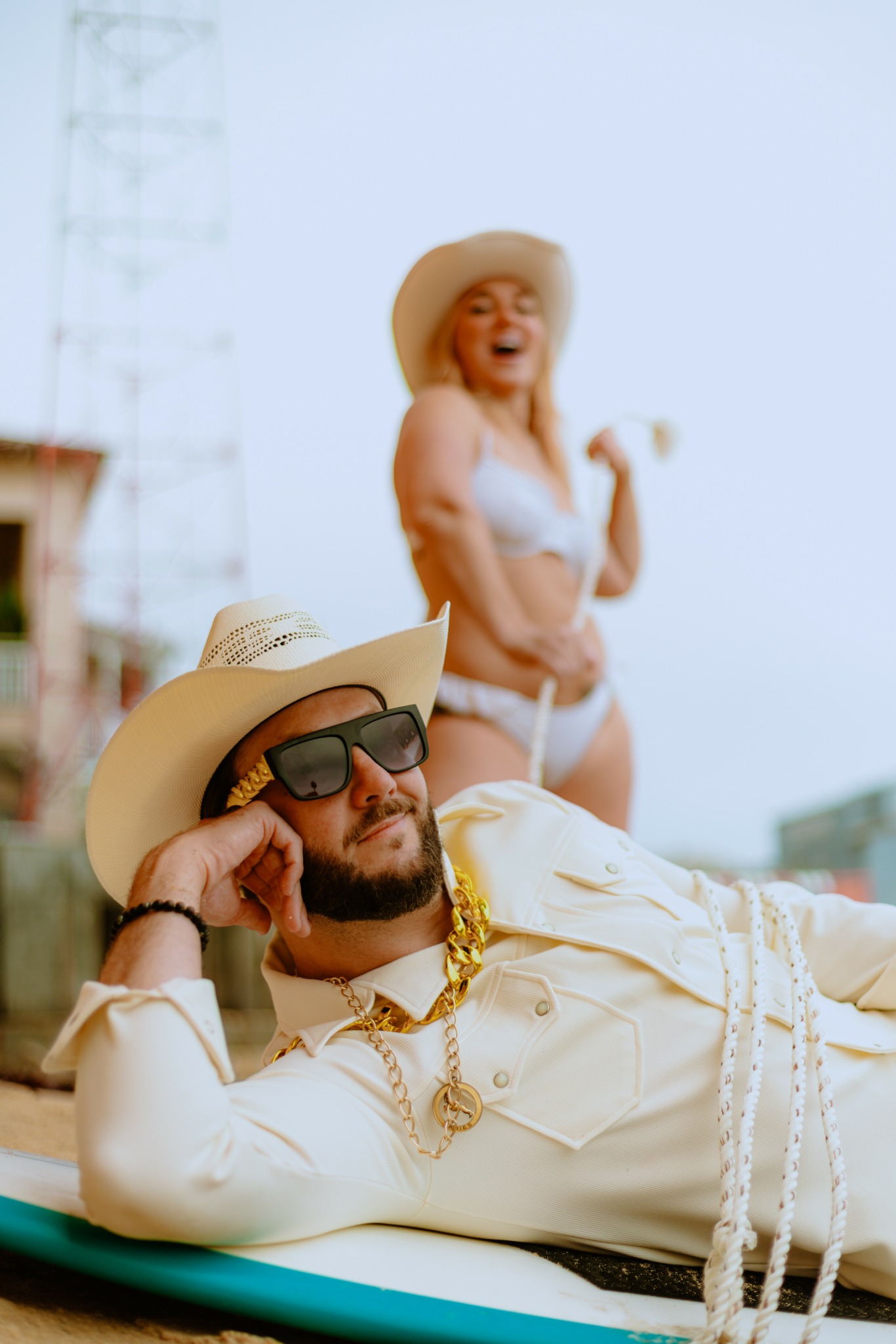 Man lying on a lounge chair wearing sunglasses, a hat, and a white outfit with gold chains, smiling. Woman in background wearing a hat and bikini, smiling.