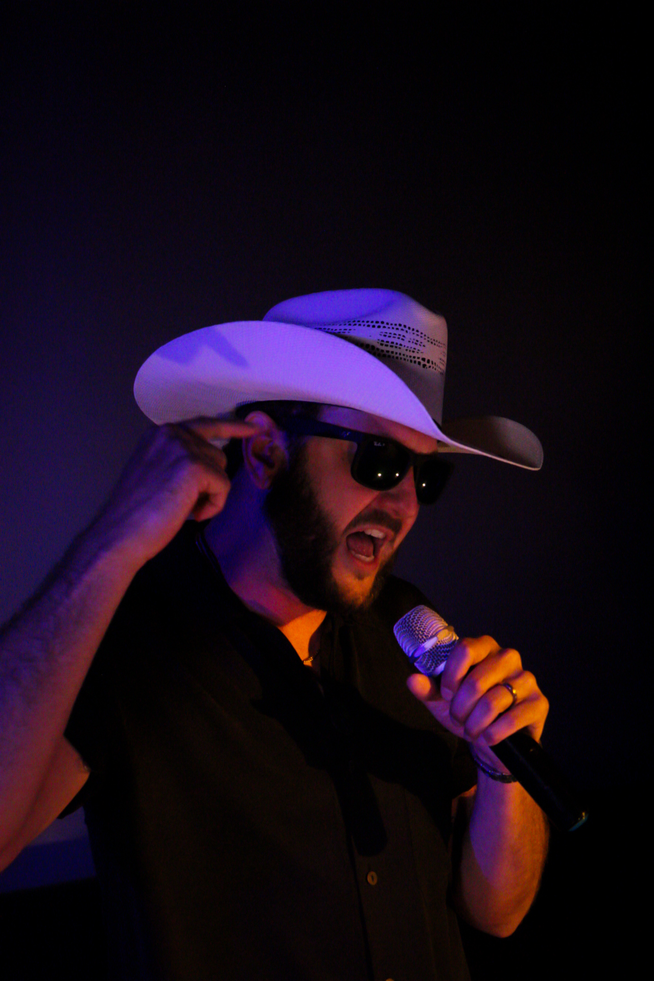 Person wearing a cowboy hat and sunglasses singing into a microphone on stage.