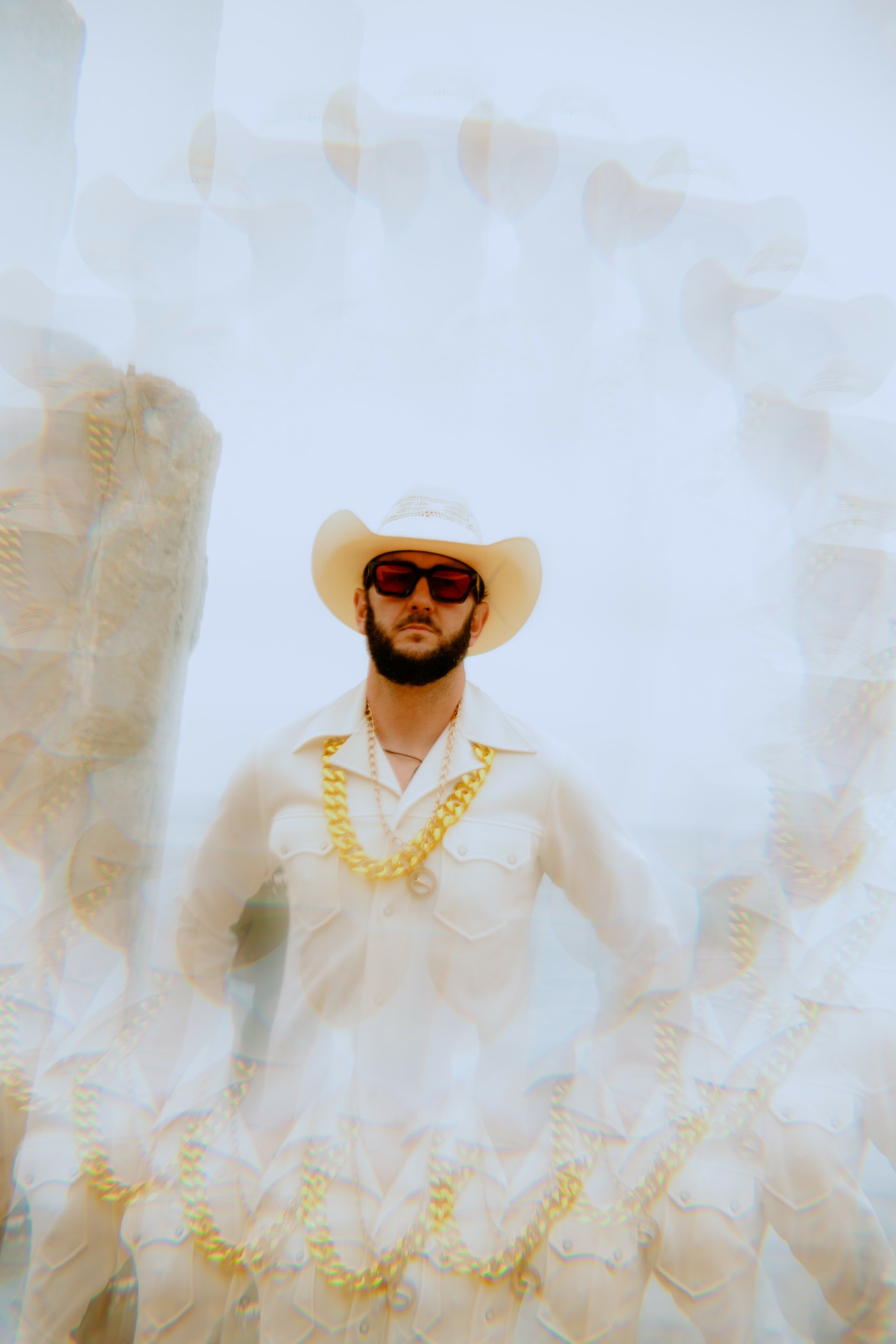 Man wearing sunglasses, a wide-brimmed hat, and a white shirt with a gold necklace, standing outdoors.