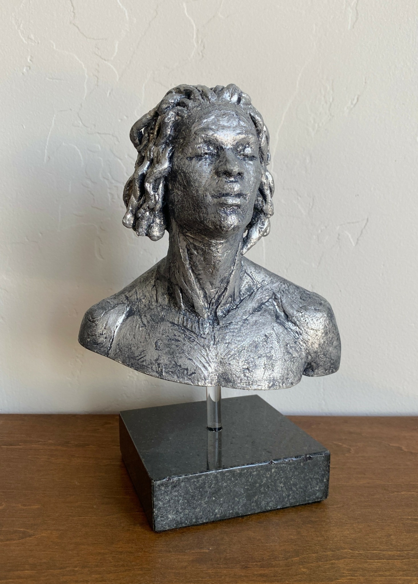 Bust sculpture of a woman with textured hair, mounted on a black rectangular base, against a plain wall.