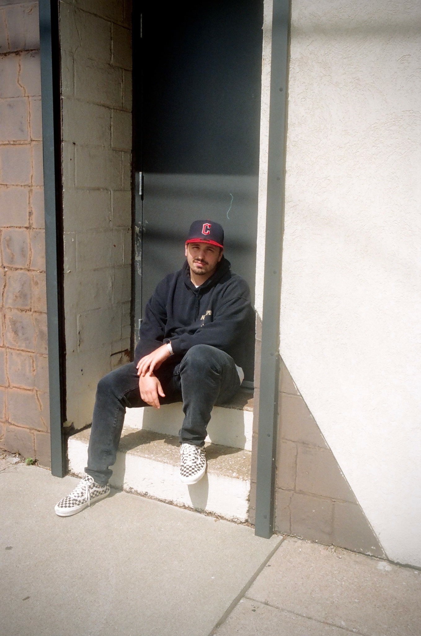 Person sitting on a step in a doorway, wearing a baseball cap, dark jacket, and sneakers, smiling.