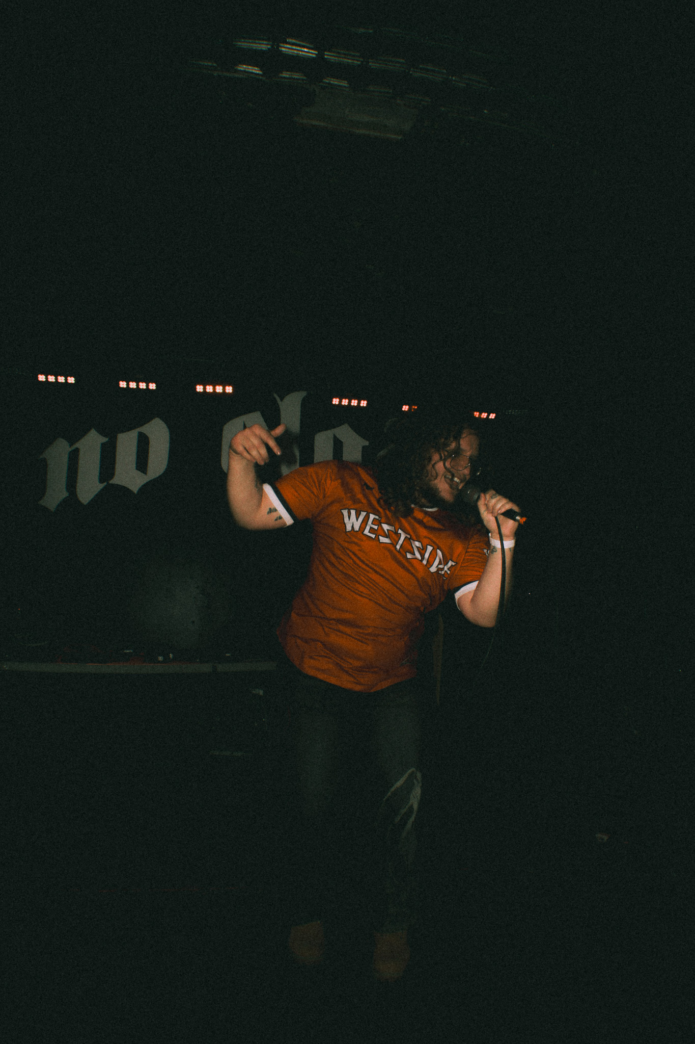 Person on stage with arms raised, wearing a red shirt with white text, in a dark environment with stage lights.