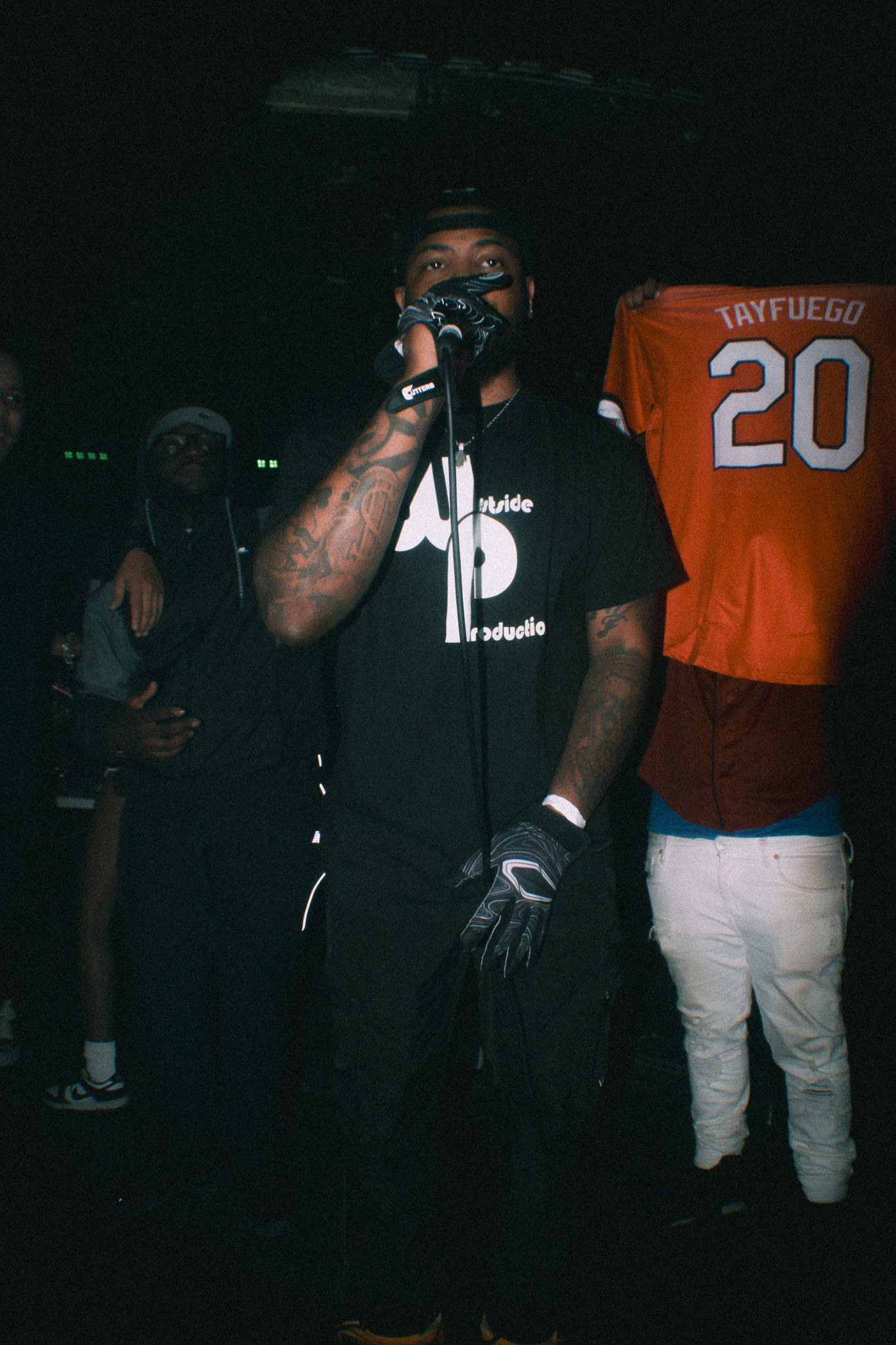 Person holding microphone on stage with others in background, dark setting, some wearing sports jerseys.