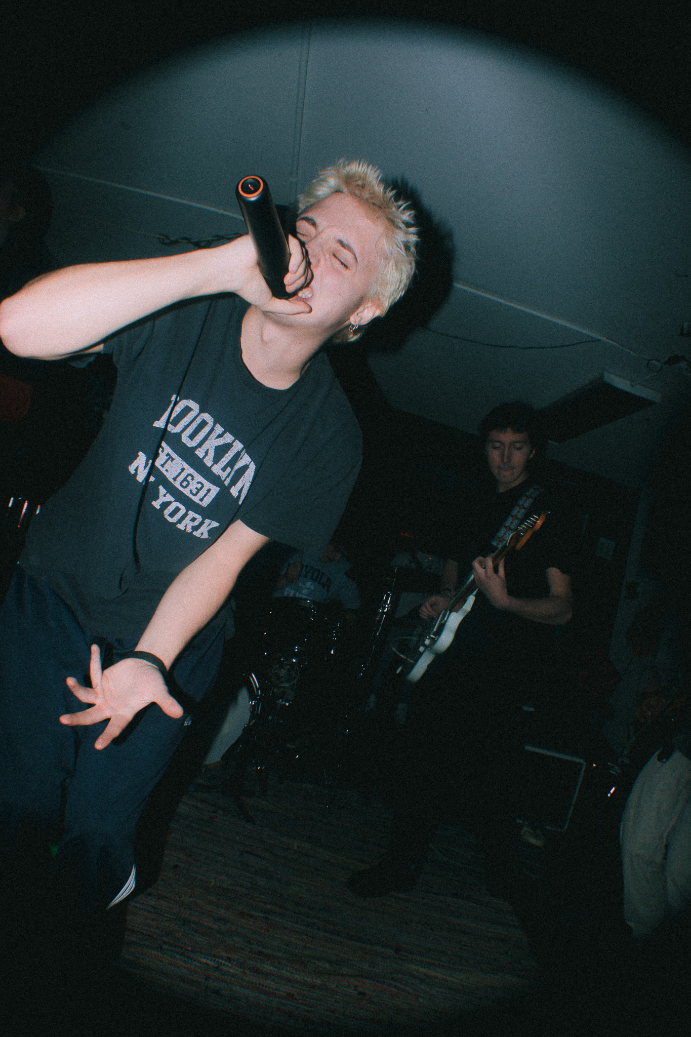 Person with short blonde hair singing into a microphone on stage, wearing a black T-shirt, in a dark room.