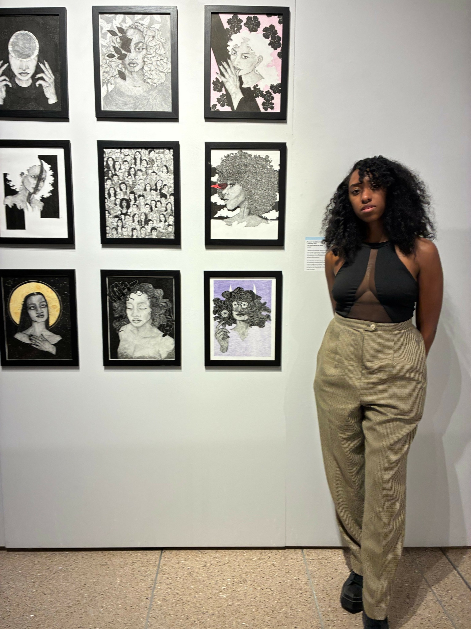 Woman with curly hair wearing black top and beige pants stands next to framed floral artwork on white wall.