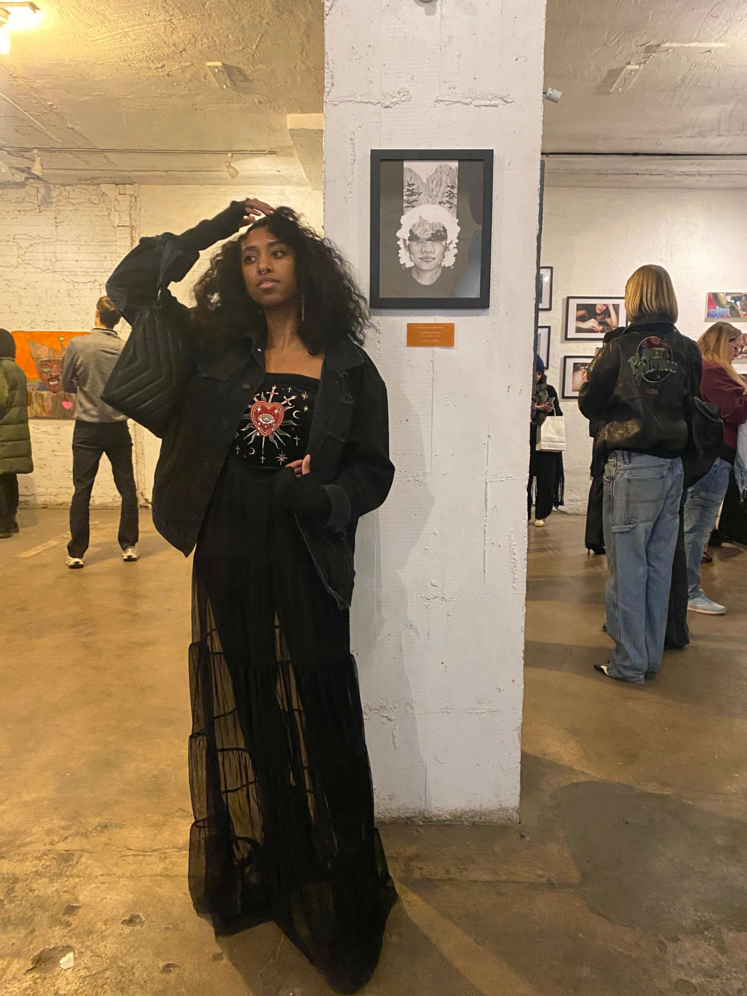 Woman in black dress and jacket standing in an indoor space, holding her hair with one hand, with people in background.