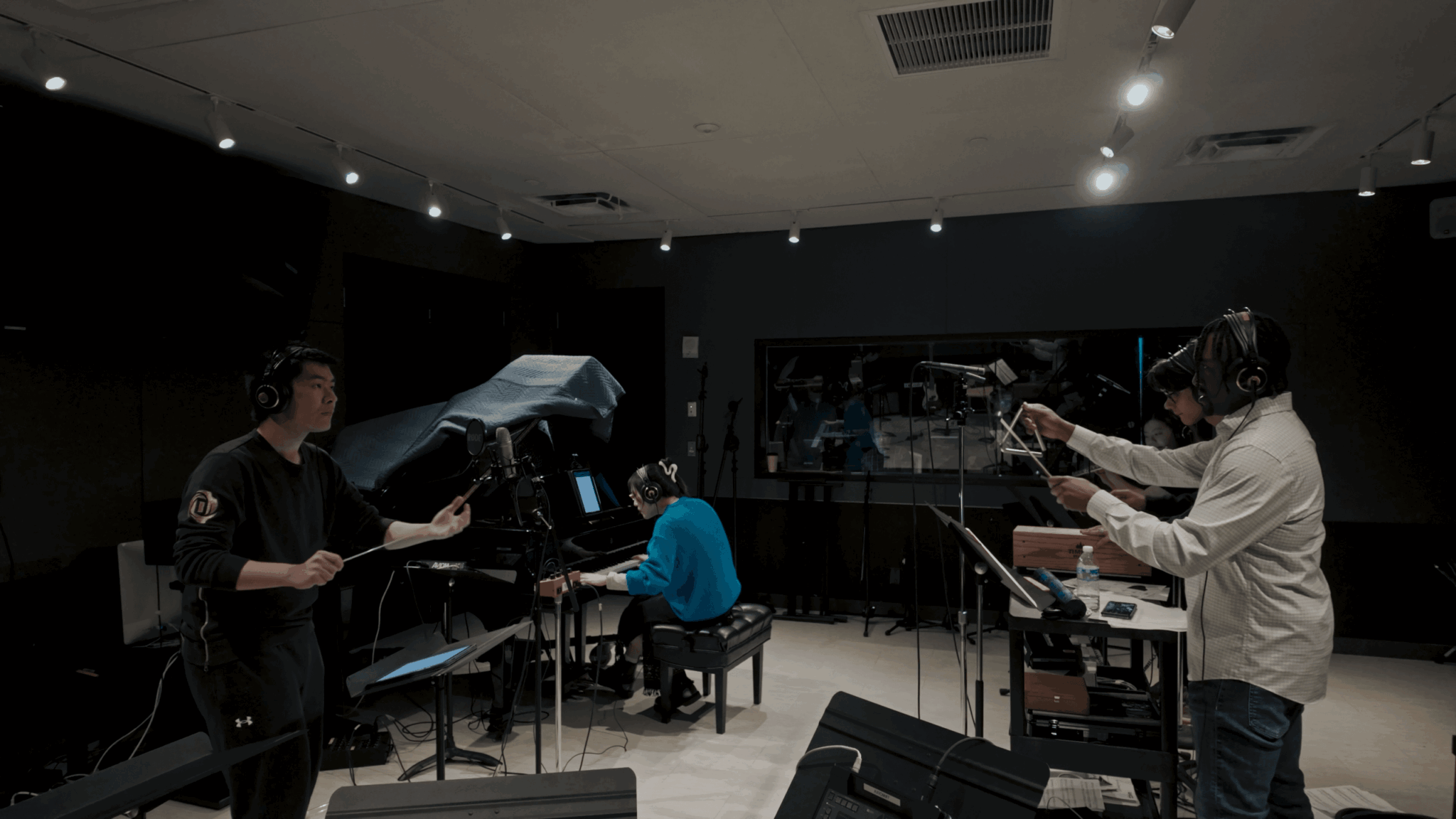 People in a recording studio with musical instruments and equipment, some reading sheet music or preparing to perform.