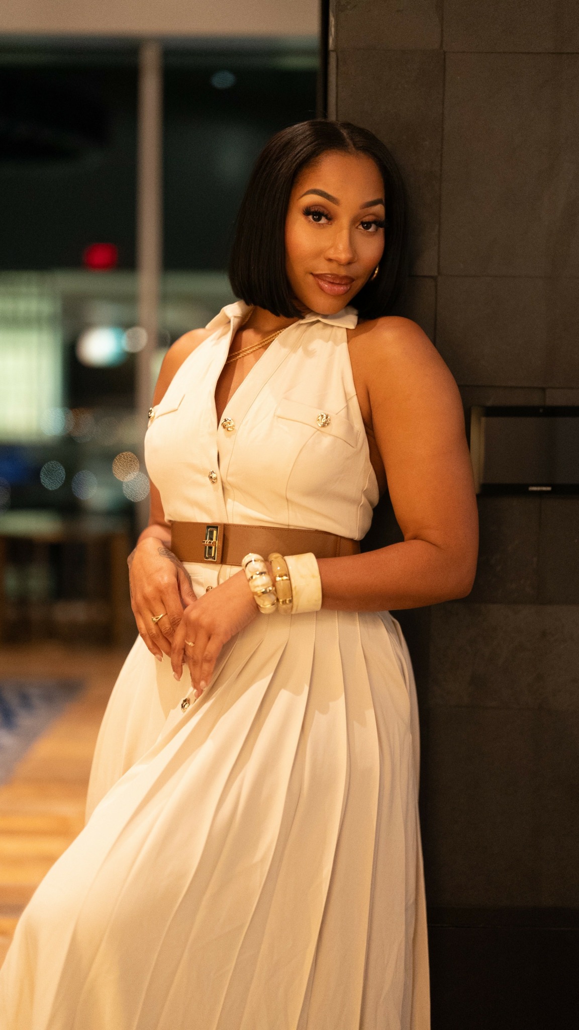Woman in white dress with brown belt and bracelets, standing indoors near a dark wall, looking at camera.