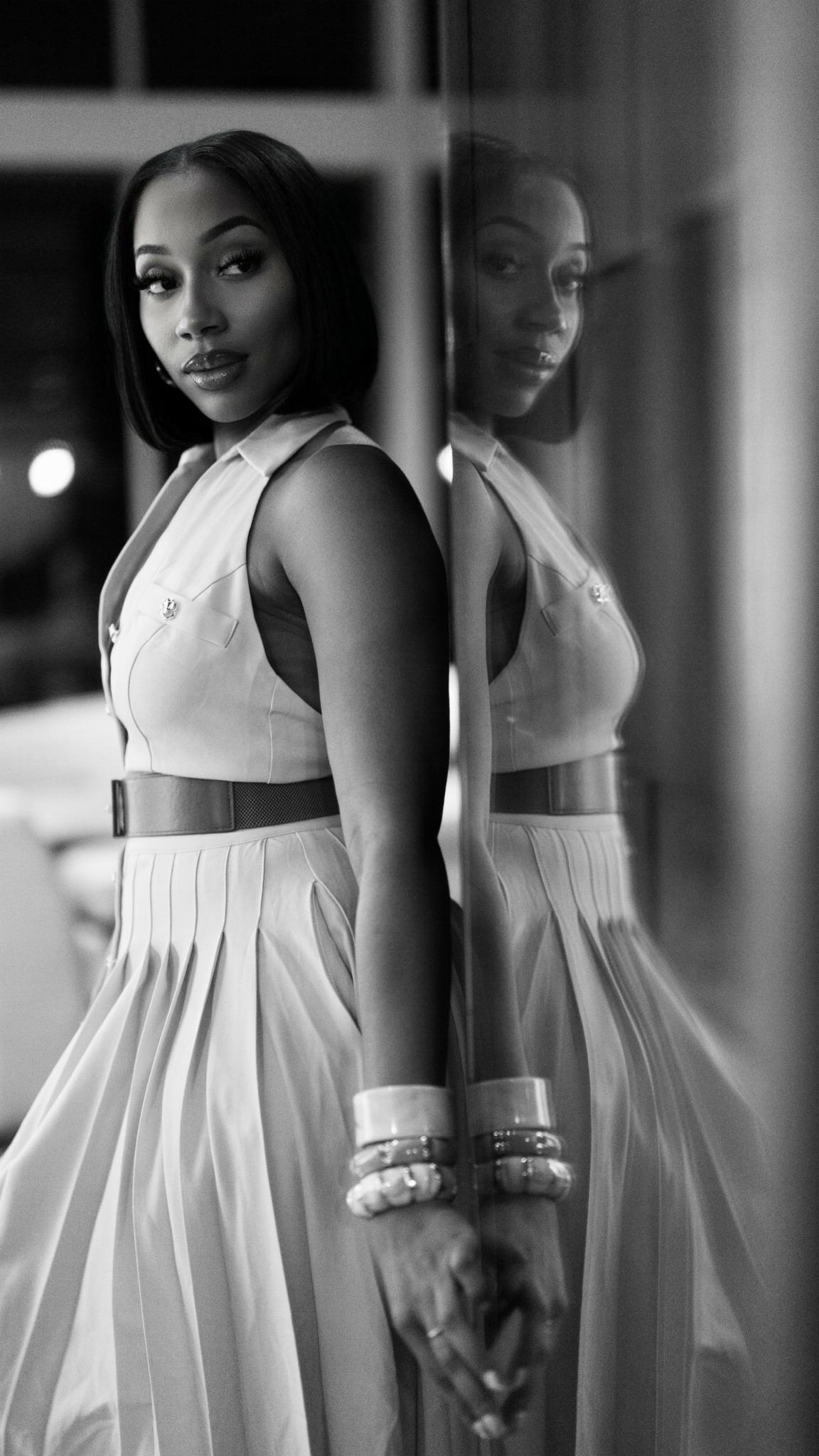 Young woman in sleeveless dress standing next to mirror, reflection visible, holding bracelets, indoors at night.