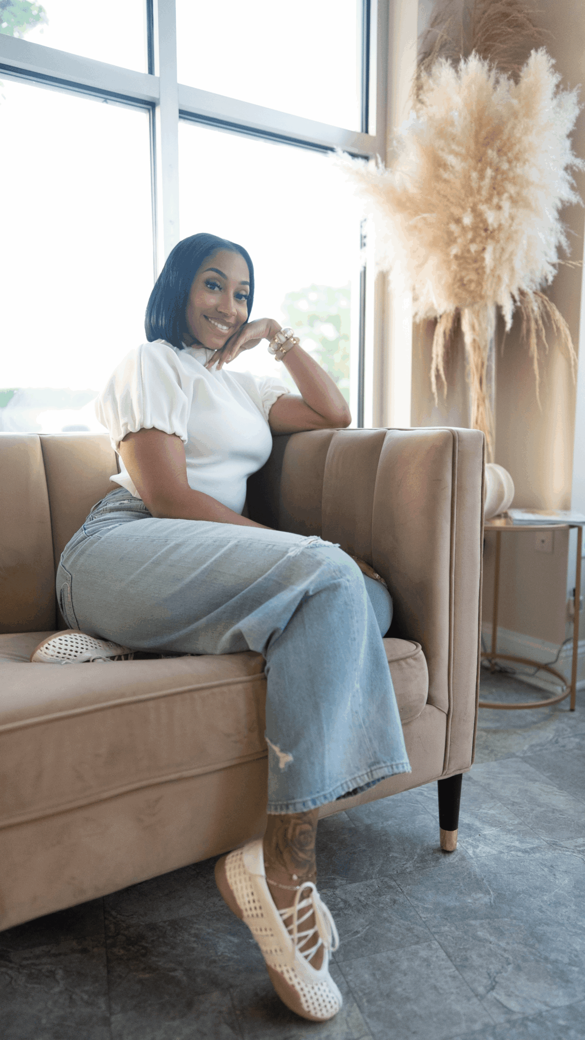 Woman sitting on sofa near window, smiling, wearing white top and jeans, with a large plant nearby.