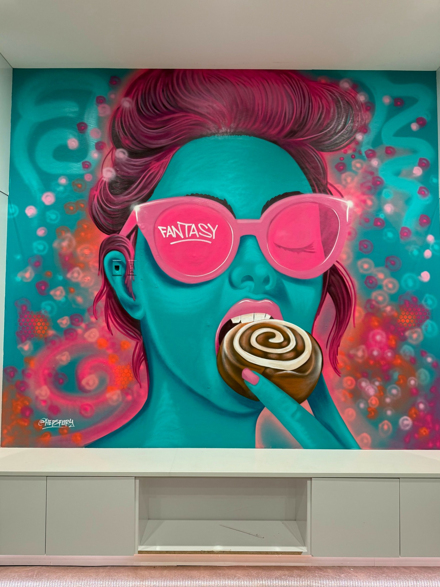 Colorful mural of a person with pink hair, wearing pink sunglasses, licking a spiral lollipop, surrounded by vibrant abstract patterns.