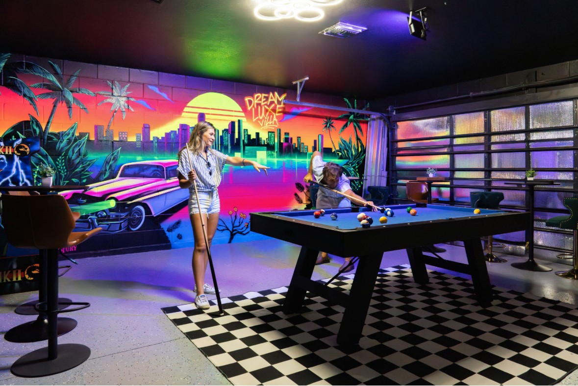 Two people playing pool in a room with a colorful cityscape mural and checkered floor.