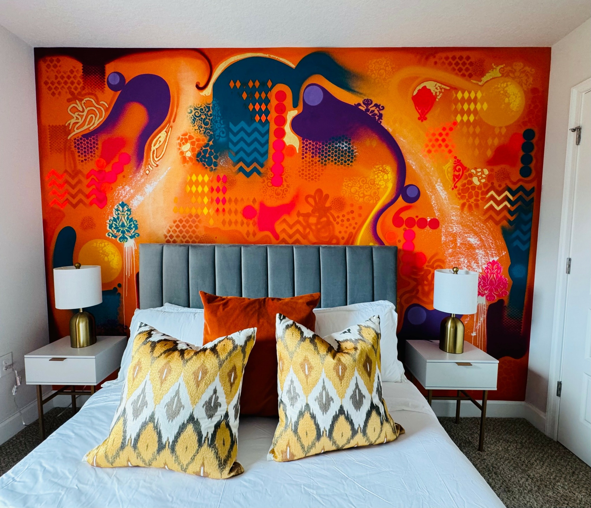 Colorful abstract mural behind a bed with gray headboard, white bedding, and decorative pillows, two white nightstands with lamps.
